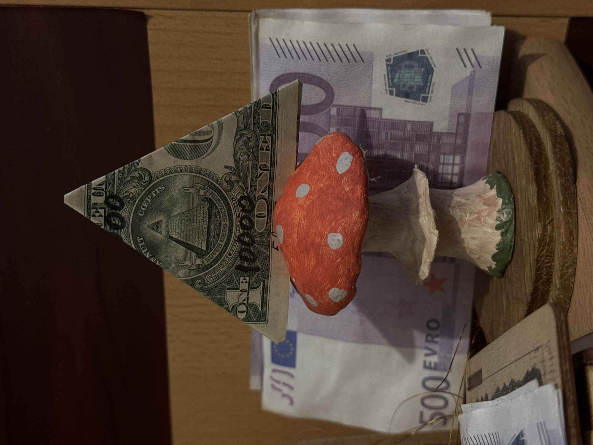 This close-up, vertically oriented image, set in Kladovitsy, Russia, displays a still life arrangement of currency and a decorative object. The main subjects are a US one-dollar bill folded into a triangular shape, a red and white spotted mushroom figurine, and several 500 Euro banknotes.
The dollar bill, positioned on the left, is folded to prominently feature the reverse side of the Great Seal of the United States, including the Eye of Providence and the Latin phrases "ANNUIT COEPTIS" and "NOVUS ORDO SECLORUM." The word "ONE" is also clearly visible. To the right of the dollar bill stands a handcrafted or decorative mushroom figurine, characterized by its vibrant red cap adorned with white spots, a light-colored stem, and a green base.
Behind these items, several 500 Euro banknotes are visible, partially obscured. The number "500" and the word "EURO" are discernible on the banknotes, along with their distinctive purple hue and security features, including a European star symbol and architectural motifs. The currency and mushroom are held within what appears to be a wooden organizer or display shelf, with light brown wooden surfaces forming the background and a darker brown surface to the far left. No activity is depicted; the scene is a static arrangement of these disparate objects.