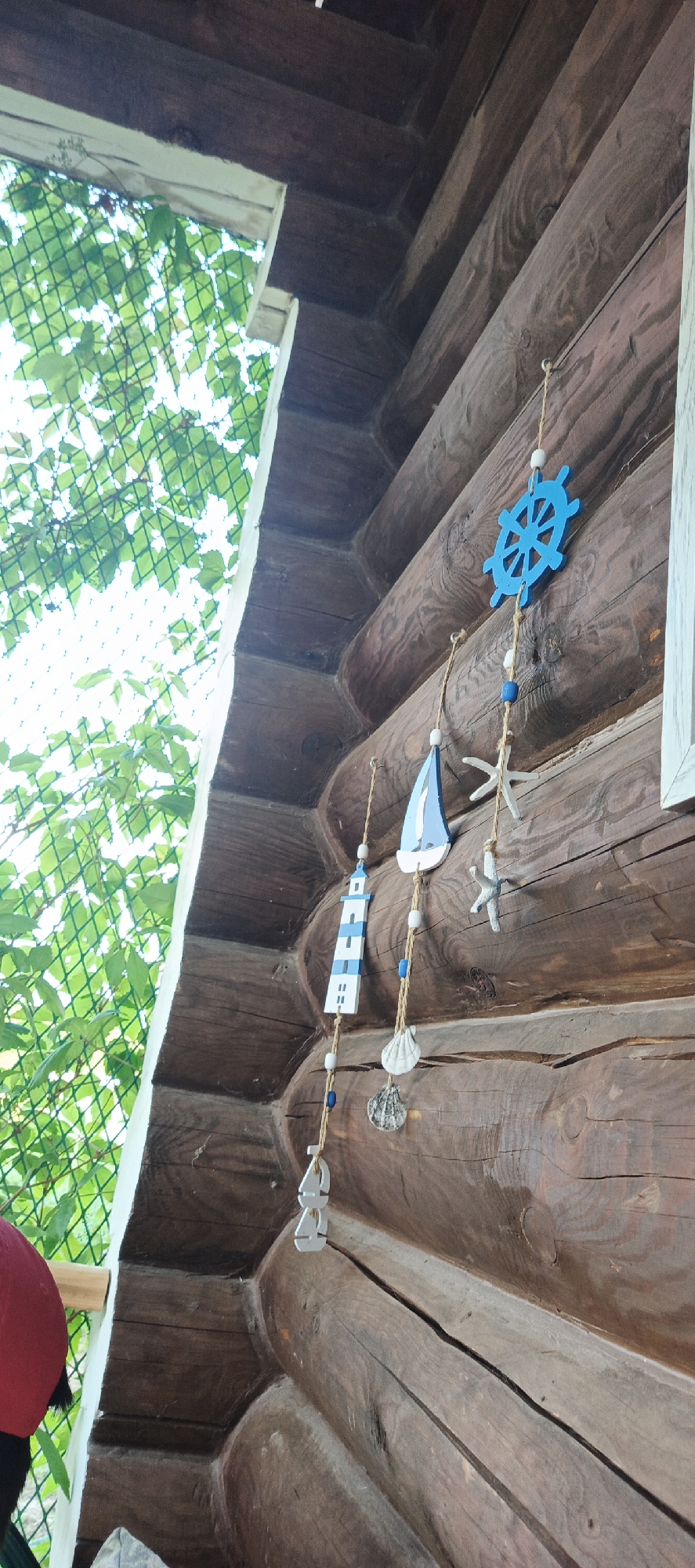 The image shows a close-up of a wooden log cabin wall, likely in Большой Букор, Russia, featuring a string of nautical-themed ornaments. The ornaments include a blue ship's wheel, a small sailboat, a lighthouse, seashells, starfish, and two abstract silver shapes. These are hung on twine with white and blue beads. To the left of the wall, lush green foliage is visible, partially obscured by a light-colored netting and a white frame, suggesting an outdoor setting such as a porch or balcony. The texture of the aged wood is prominent, with visible grain and some natural cracks. In the bottom left corner, a small portion of a person wearing a red cap is visible.