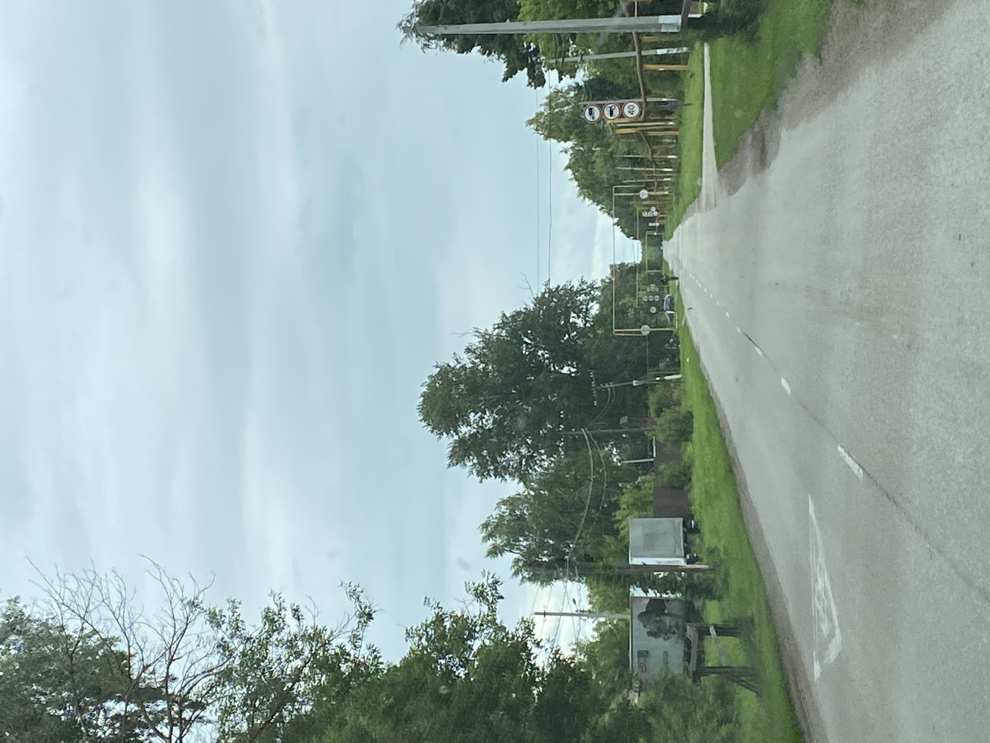The image depicts a straight, paved road leading into the distance, lined with trees and vegetation on both sides. The sky is overcast with a pale blue hue. On the left side of the road, there are some trailers and a wooden structure. On the right side, there are multiple traffic signs, including a speed limit sign indicating 40. A single person is walking on the road in the distance. The scene appears to be a rural or suburban area. The location is identified as Nadezhdino, Russia.