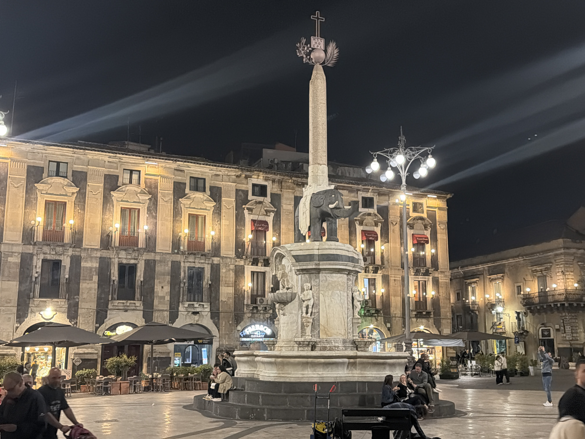 The image shows Piazza del Duomo, Catania, Italy, at night. The Fontana dell'Elefante dominates the center, featuring an elephant statue supporting an Egyptian obelisk topped with a cross. People are sitting around the fountain's base, while others are walking in the plaza. Behind the fountain, buildings with lighted windows rise, including a facade with awnings and lights over the windows. There are outdoor cafe umbrellas and seating around the plaza. Overhead, lights streak across the night sky.
