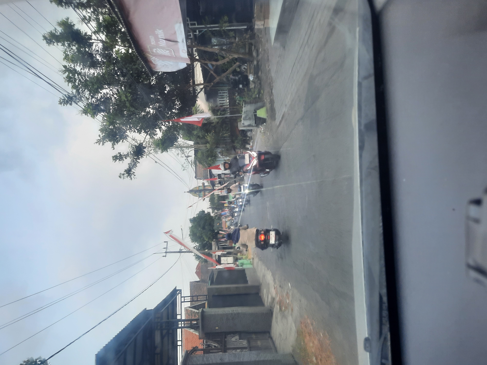 The image depicts a street scene in Banjarkemantren, Indonesia, with several individuals riding motorcycles. The street is paved and lined with houses and trees. Indonesian flags are visible, indicating a possible national celebration or event. The perspective suggests the photo was taken from inside a vehicle. The sky is overcast.