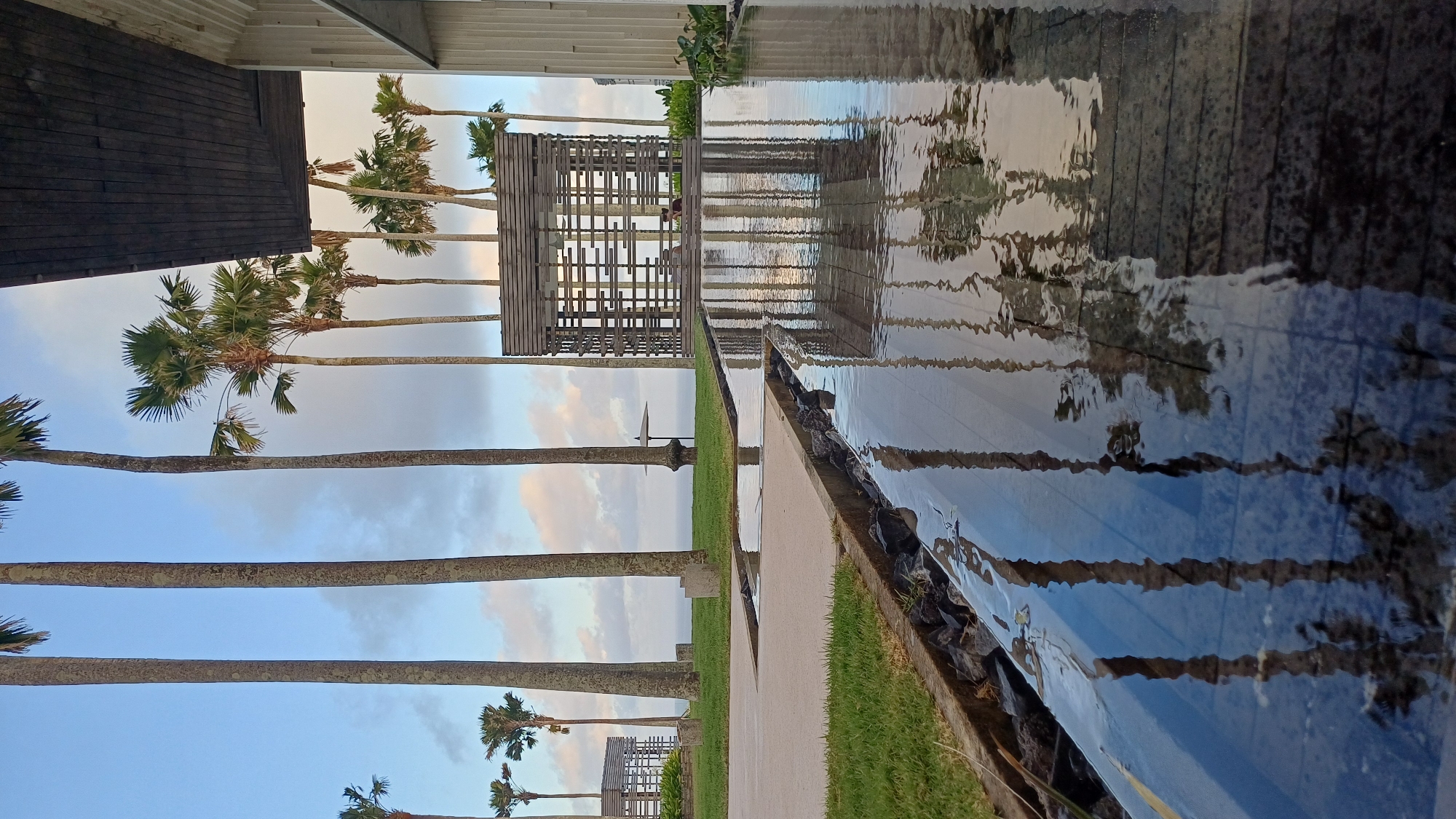 The image shows an outdoor scene in Kuta Selatan, Indonesia. There are palm trees lining a walkway with a water feature on the right. The water feature reflects the sky, palm trees, and a wooden structure in the distance. The walkway is lined with grass and rocks. A building is partially visible on the left side of the image. The wooden structure is a shaded area with seating. There is a person sitting in the shaded area. The sky is cloudy with a mix of blue and white.