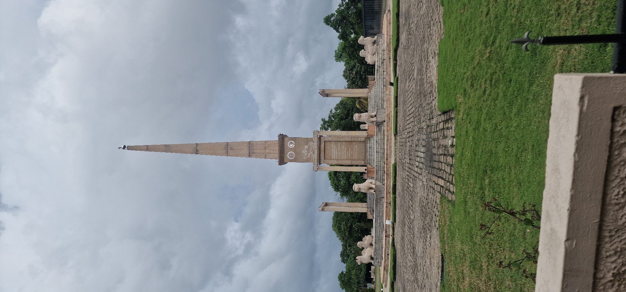 The image shows the Sri Jayawardenepura Kotte National Monument in Sri Lanka. The monument consists of a tall, tapering obelisk with two circular carvings in the upper section, supported by pillars and surrounded by lion statues. The monument is located in a park-like setting with green grass and trees. The sky is overcast with grey clouds. A stone walkway leads to the monument. A section of a building wall or another part of the monument is visible in the lower right corner of the image.