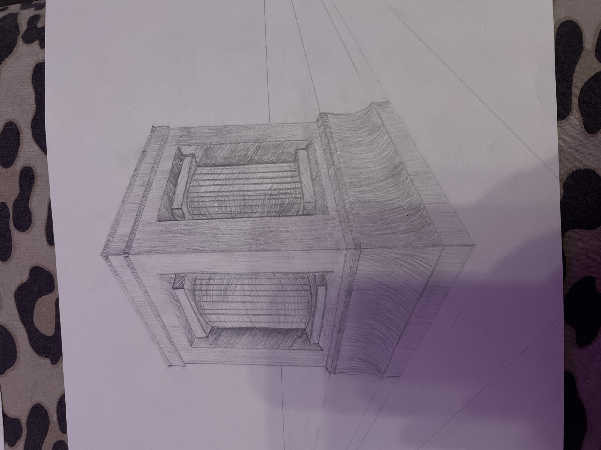 The image is a pencil drawing on paper, depicting a classical architectural element, possibly a monument or a pedestal. The structure is presented in a perspective view, suggesting it is placed on a flat surface that recedes into the distance, indicated by drawn lines. The main subject is a rectangular base topped with a more intricate section featuring recessed columns. The drawing exhibits shading and line work to create depth and texture, particularly on the fluted columns and the curved base. The background is plain white paper, and the image appears to be taken indoors, with a patterned fabric visible at the top and bottom edges. There are no people or animals in the drawing, nor any specific urban context. The location in Zmeinyi, Russia, is not depicted or suggested by any elements within the artwork.