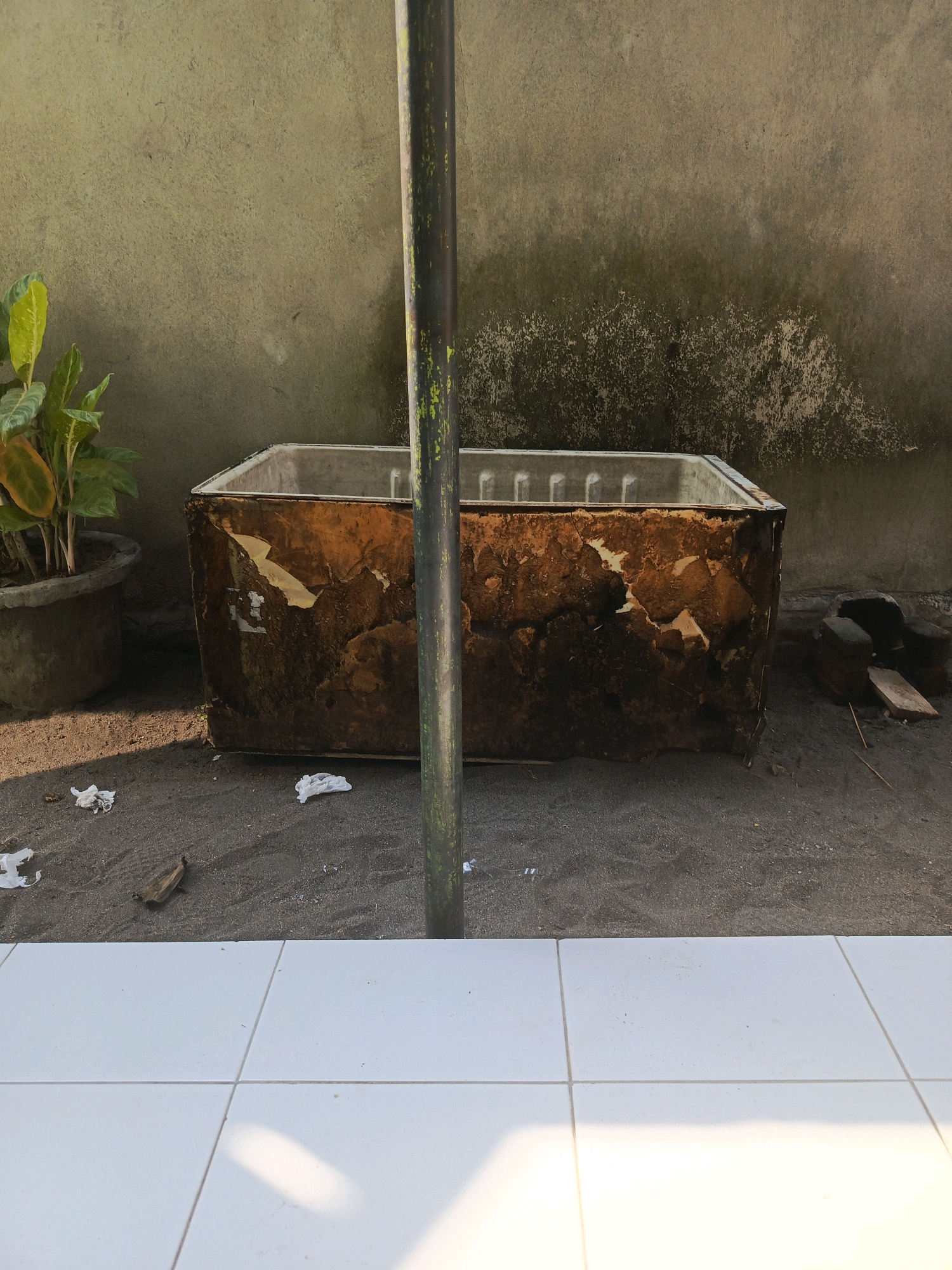 A close-up, slightly high-angle shot captures a weathered, rectangular bathtub resting on dark sand. The bathtub's exterior is heavily damaged, with peeling brown and yellow material revealing a rougher surface beneath. To the left, a potted plant with green and yellow leaves stands beside the tub. In the foreground, a section of white tiled flooring occupies the bottom portion of the image. A vertical, dark pole with greenish streaks runs through the center of the frame, partially obscuring the bathtub. The background features a textured, pale greenish-gray wall with visible signs of age and discoloration. Scattered white paper or plastic debris is present on the sand. The scene appears to be outdoors, possibly in a backyard or an open area within the city of Lubuklinggau, Indonesia, suggesting a scene of disuse or abandonment.