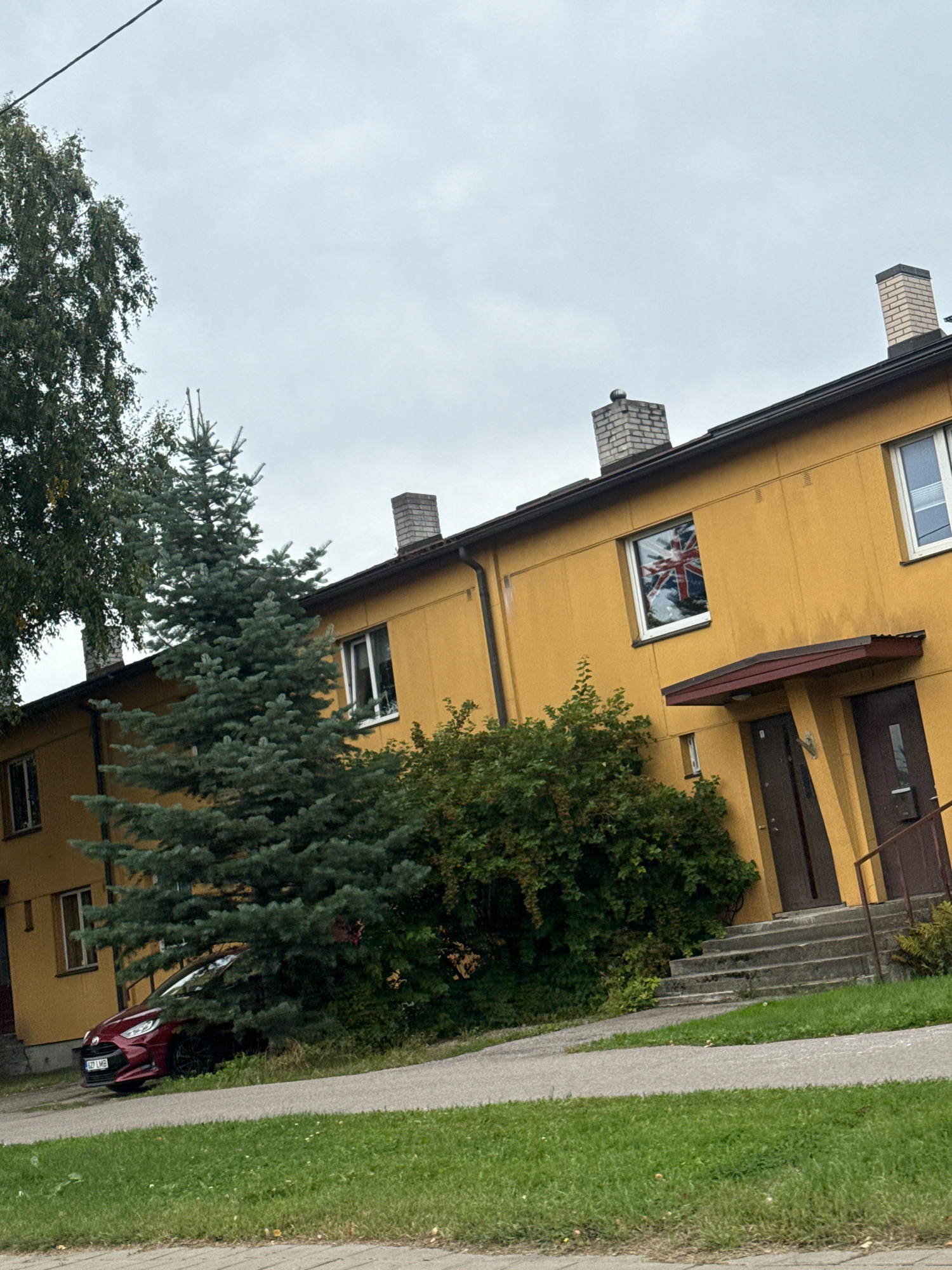 The image depicts a view of a row of yellow apartment buildings in Tallinn, Estonia. In front of the buildings, a dark red hatchback car is parked under a large coniferous tree, partially obscured by foliage. The car's license plate reads "627 LMB". A paved walkway runs in front of the car and continues past the buildings. To the right, concrete steps lead up to two entrances with dark brown doors and burgundy awnings. One of the windows on the second floor of the building reflects the Union Jack flag. The sky is overcast and grey. Green grass covers the foreground and areas around the buildings.