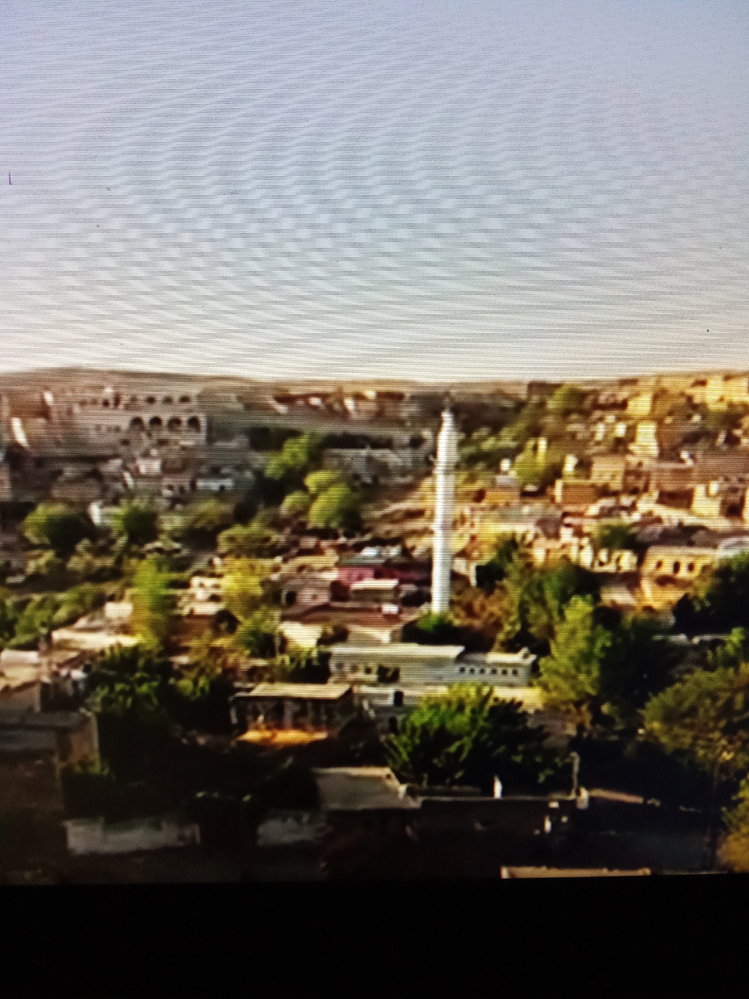 The image displays an aerial view of a village with a prominent minaret, suggesting a location in a region with Islamic architecture. The village consists of numerous buildings with terracotta roofs, interspersed with green trees and vegetation. The structures appear to be made of stone or mud brick, and the overall landscape is hilly and arid. The sky is a light blue with horizontal lines indicating a digital display or screen artifact. There are no visible people or animals in the image. The scene is characterized by its rustic and traditional architecture, typical of a rural settlement in Central Asia. Without further context or more distinct visual cues, pinpointing the exact location within Рощин ауылдық округі, Kazakhstan is not possible.