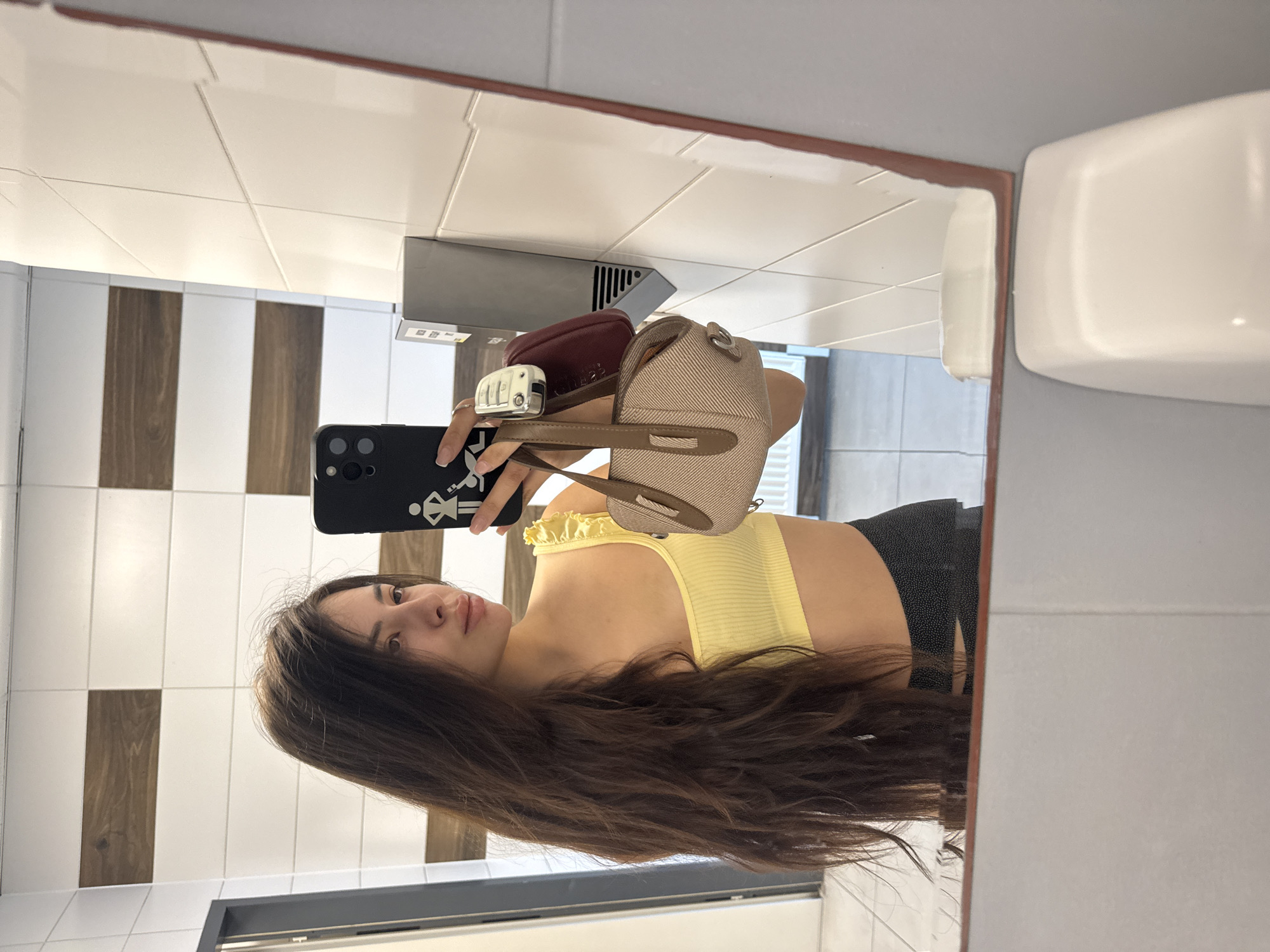The image is a mirror selfie taken by a young woman in what appears to be a public restroom or changing room. The woman, who has long, dark brown hair cascading over her shoulders, is wearing a ribbed, bright yellow crop top with ruffled detailing at the neckline and dark-colored bottoms. She is looking directly towards the camera with a neutral expression.
She is holding a black smartphone in her left hand, which has a distinctive white graphic on its case depicting stick figures of a person in a dress and another in pants, possibly representing restroom symbols. Attached to the phone, and held in her hand, is a car key with a silver casing, and a small, beige, textured bag with a brown strap. This bag seems to be resting on or attached to her hand/wrist, close to the phone.
The background, as reflected in the mirror, features white square tiles on the upper wall and alternating white and dark brown, wood-patterned vertical rectangular tiles on the lower half. Above her hand, a silver, wall-mounted fixture, likely a hand dryer or dispenser with a vent, is visible. To the far right, a large white, rounded fixture, possibly a soap dispenser or another part of a restroom amenity, is mounted on the wall. The mirror itself has a reddish-brown frame along the top and right edges. The location context for this scene is Plovdiv, Bulgaria.