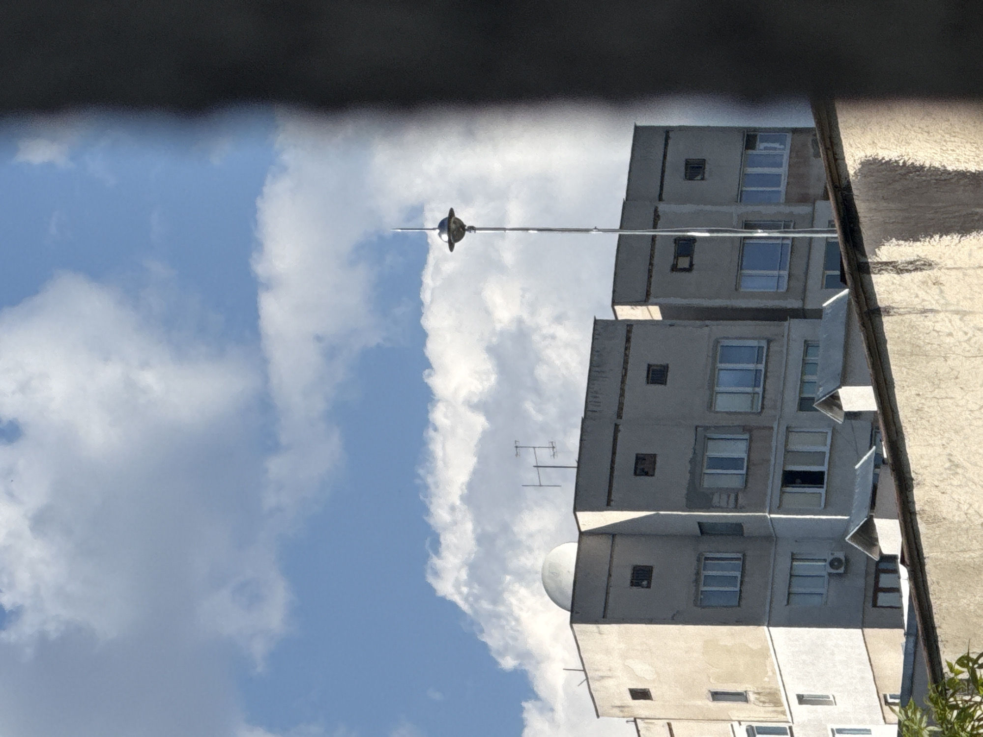 A low-angle shot looks up at a tall, light-colored building against a backdrop of blue sky filled with white clouds. The building appears to be a multi-story residential structure, with numerous windows visible on each floor. Some windows have air conditioning units installed. An antenna extends from the top of the building, reaching into the sky. A dark, indistinct object obscures the view along the top edge of the frame, possibly part of a structure the photo was taken from. A light-colored wall or structure is visible on the right side of the frame, with some greenery at the bottom corner.