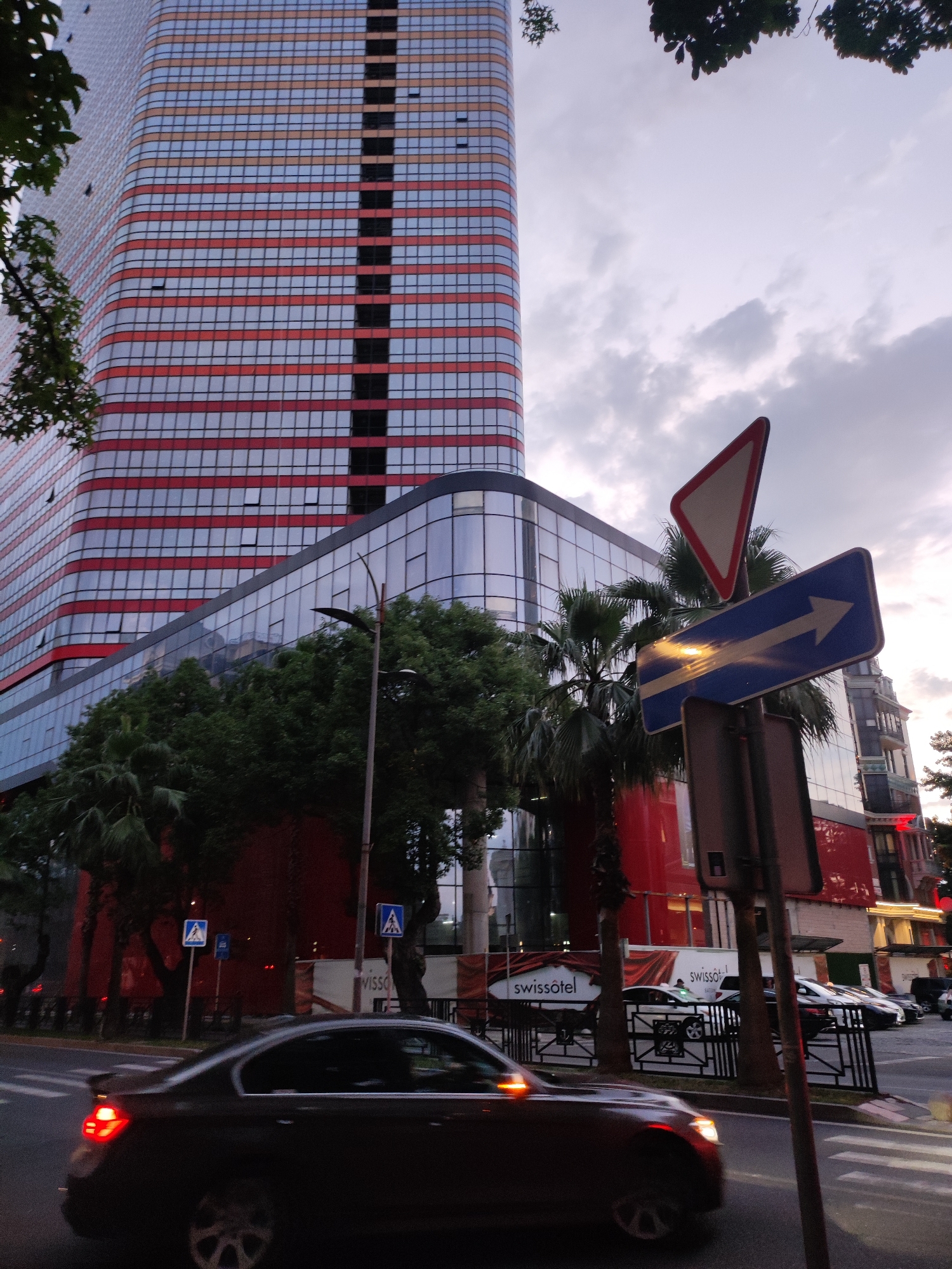 The image captures a street scene in Batumi, Georgia, featuring a modern skyscraper with a distinctive red and white striped facade. The skyscraper has large, reflective glass windows. In the foreground, a dark-colored sedan is in motion on a street, with its brake lights illuminated. Traffic signs are visible, including a yield sign and a blue directional sign with a white arrow pointing to the right. Palm trees and other foliage are present, interspersed with the buildings. A lamppost stands on the right side of the street. In the background, other buildings with intricate architectural details and illuminated windows can be seen. A banner with the text "swissôtel" is visible on the side of one of the buildings. The sky is overcast with some clouds. The overall impression is of a city environment during twilight or an overcast day.