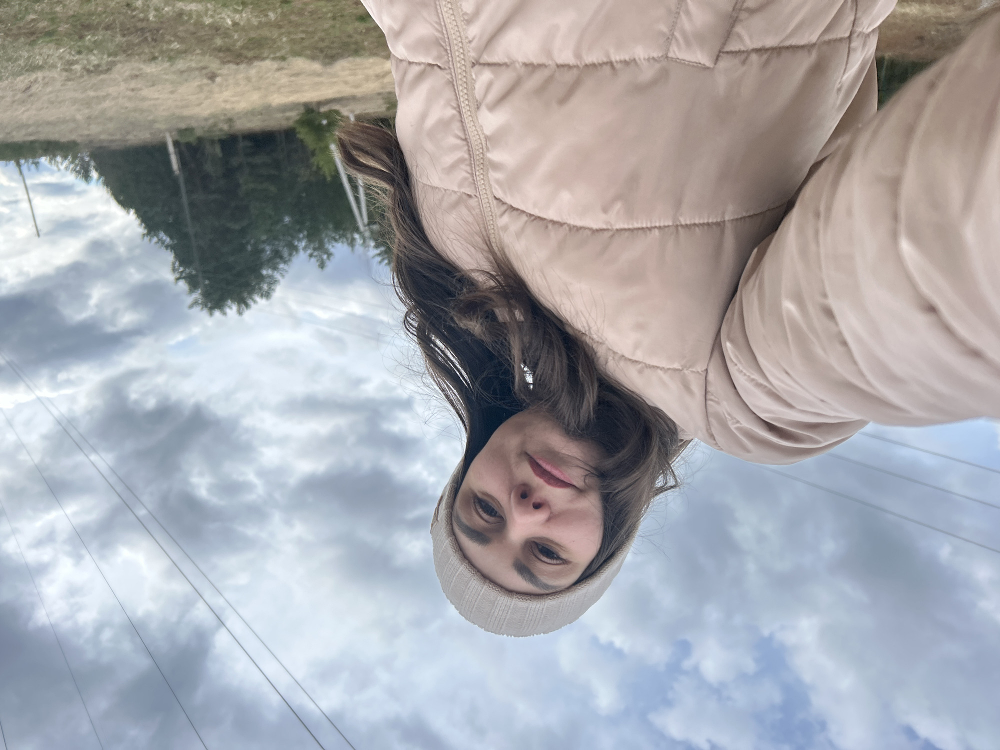 The image, originally rotated, depicts a young woman taking a selfie outdoors in what appears to be a rural or semi-rural setting in Фоки, Russia. The woman is positioned centrally, looking directly at the camera with a neutral and calm expression. She has long, dark brown hair, and is wearing a light-colored (beige or tan) ribbed beanie and a matching puffy winter jacket, suggesting cool weather.
The background features a broad expanse of overcast sky filled with grey and white clouds. Below the cloud line, several thin, dark power lines or utility wires stretch across the frame. Further back, on the horizon, there's a patch of dry, light-colored ground or grass, behind which a dense cluster of dark green evergreen trees forms a forest or wooded area. A few faint vertical poles, possibly utility poles, are visible within the tree line.
The activity taking place is a self-portrait (selfie) session. The time of day is daytime, but the heavy cloud cover prevents clear sunlight, indicating an overcast day. No specific event is evident beyond the selfie. There is no visible text in the image. The overall impression is one of a quiet, possibly cool, outdoor moment.