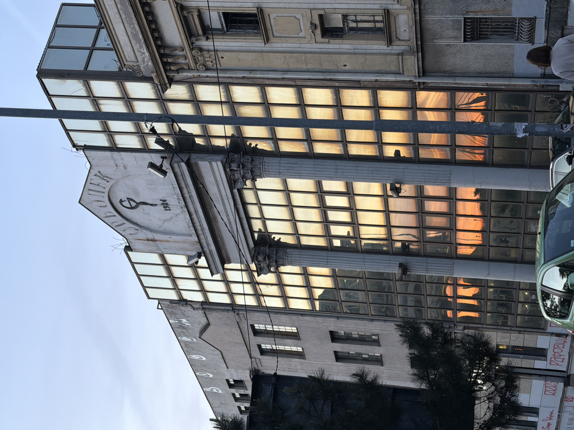 The image depicts a building facade in Belgrade, Serbia. The building has a large glass-covered section reflecting the orange hues of a sunset. A prominent detail is the inscription "АПОТЕКА 1924" on the facade, indicating it is a pharmacy established in that year. Architectural elements include columns with ornate capitals. A street light pole obscures part of the view. A car is partially visible. The surrounding buildings have a more traditional architectural style with rectangular windows. Graffiti is on the side of the building.