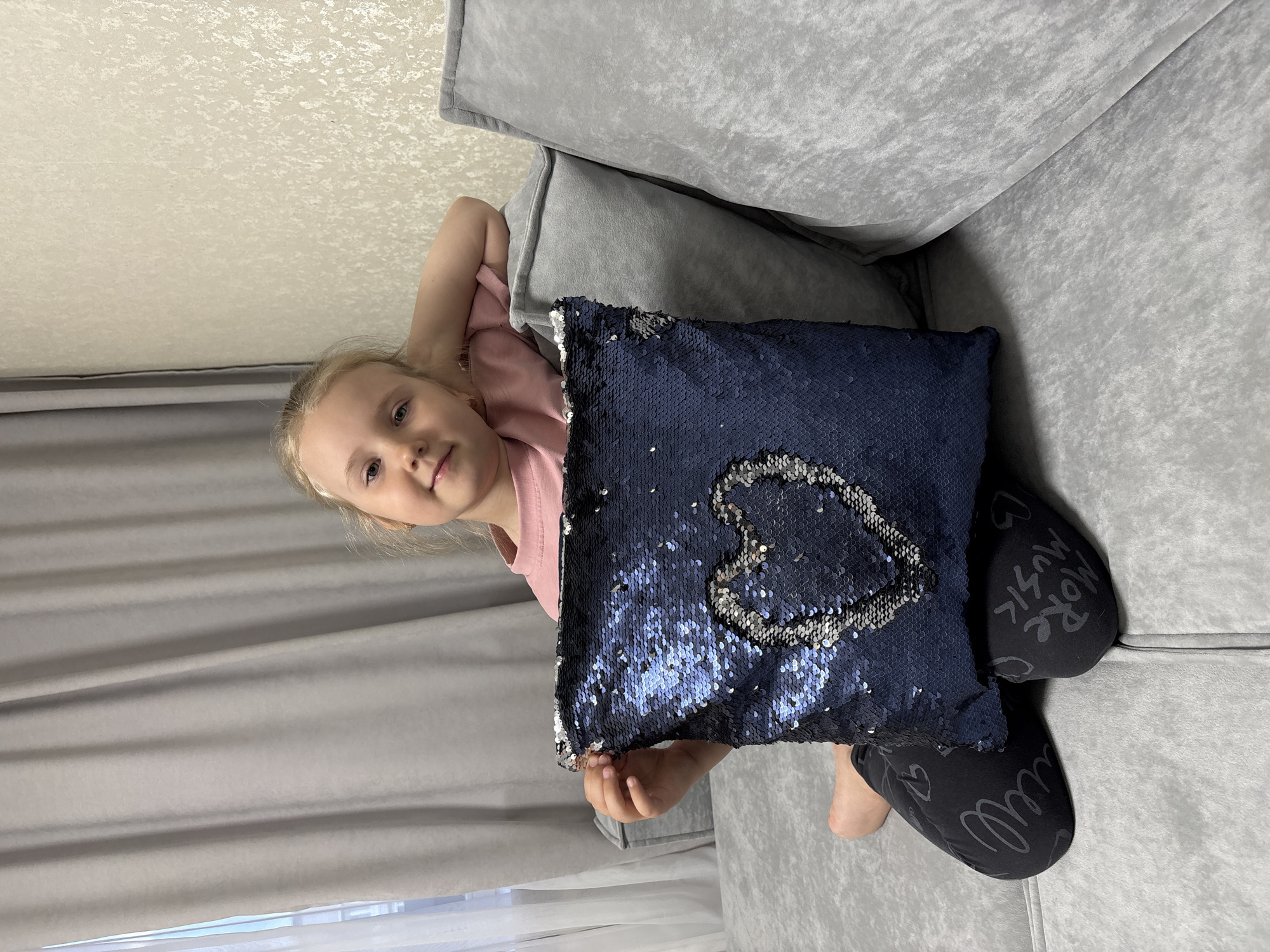 A young girl lies on a gray couch, holding a blue sequined pillow with a silver heart design. She is wearing a pink shirt and black leggings with the word "MUSIC" printed on them. The scene is likely indoors, with a light-colored wall and gray curtains visible in the background. The location is Chaikovsky, Russia.