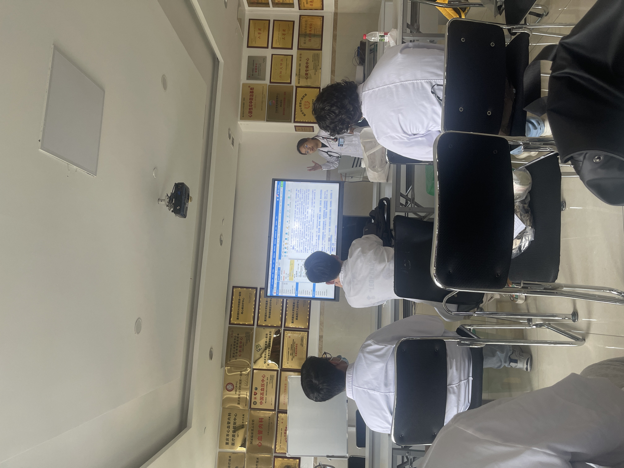 The image shows a medical conference or training session in a room filled with numerous award plaques displayed on the wall. Several individuals wearing white medical coats are seated and facing a presenter who is gesturing towards a large screen displaying a complex document or spreadsheet. The session appears to be taking place in a clinical or institutional setting. The awards on the wall indicate the location is in Yuzhong District, China.