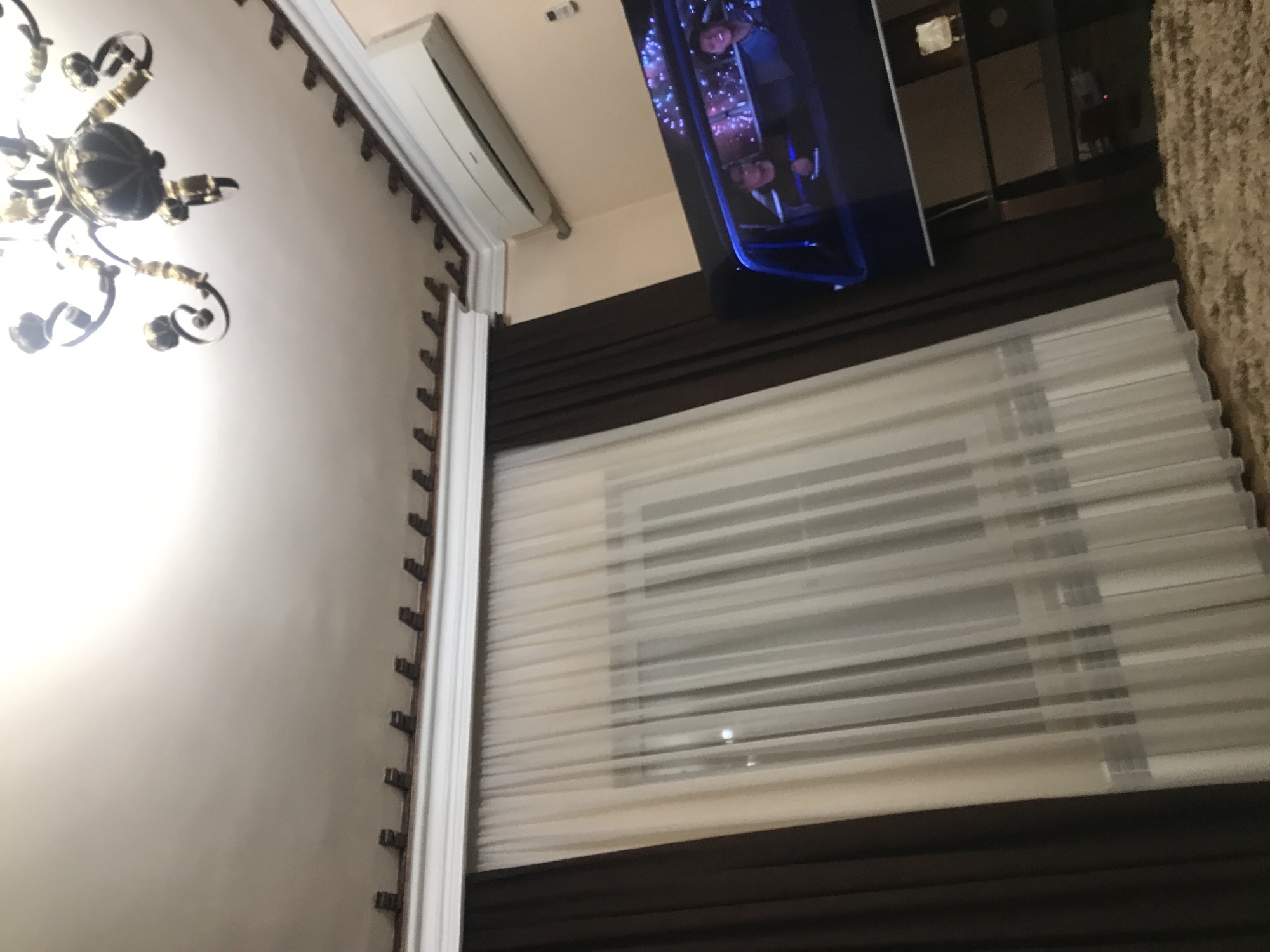 The interior shot shows a room with a chandelier hanging from the ceiling on the left side. A window covered with layered blinds and dark drapes is visible on the right side. A television is mounted on the wall above the window. The photo does not provide sufficient information to identify the location in Yerevan, Armenia, or any details about people, subjects, or interactions.