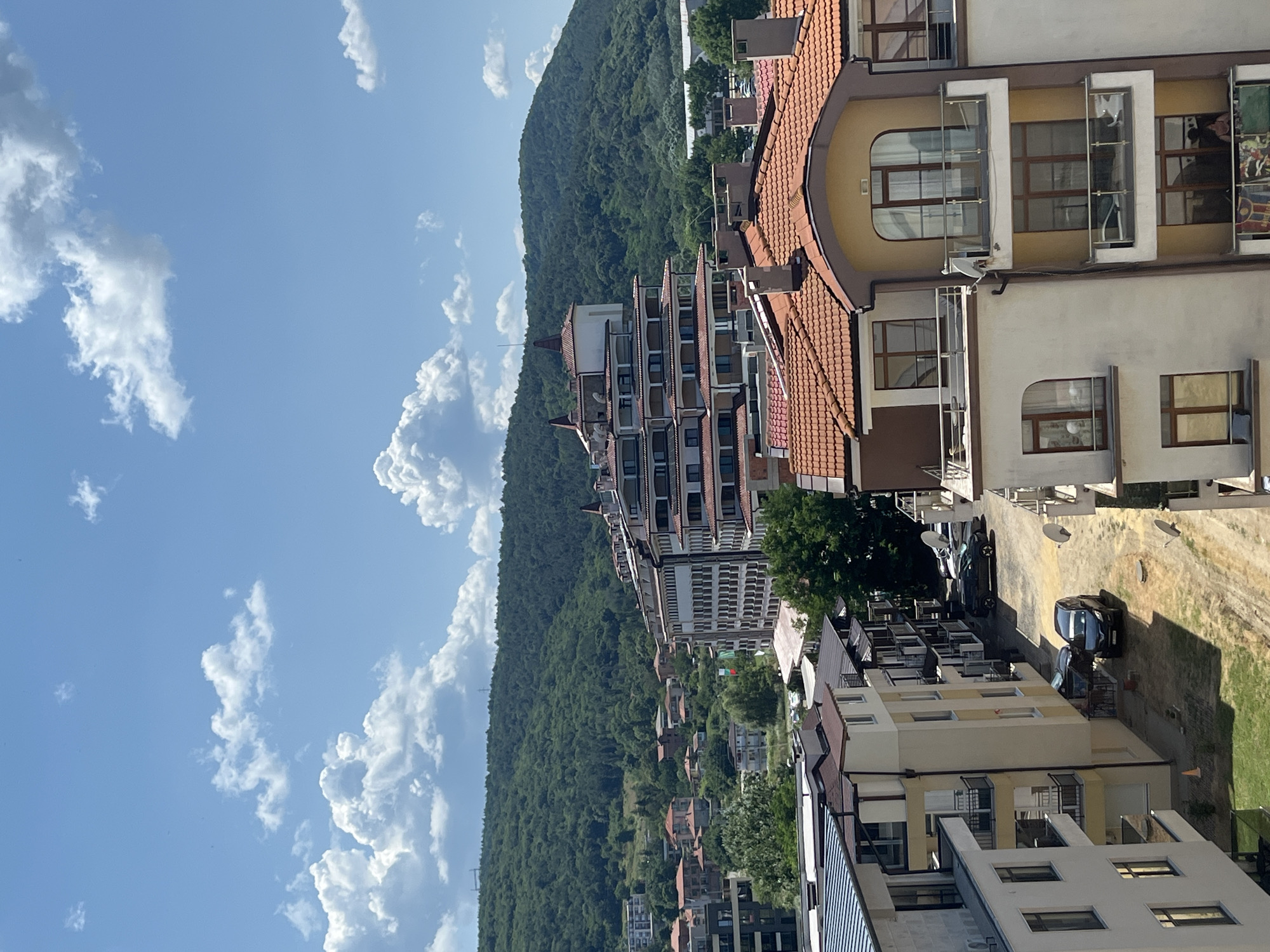 This is a drone shot taken from above in Obzor, Bulgaria, showcasing a residential area nestled at the foot of a lush, green hill. In the foreground, the view is dominated by the rooftops and upper floors of several apartment buildings, characterized by their red-tiled roofs and cream-colored facades. Balconies with glass railings are visible, along with various architectural details like arched windows and chimneys. The buildings are densely packed, creating a cohesive urban landscape.
Behind these structures, a road winds through the neighborhood, with several cars parked along its sides, including a dark-colored van and SUV. Scattered trees and smaller residential houses with similar red roofs are interspersed throughout the scene, adding to the residential feel.
Further in the background, the prominent feature is a large, terraced hotel or apartment complex with a distinctive stepped design and red-tiled roofs, situated closer to the hill. Behind this complex, a steep, densely forested hill rises, covered in a vibrant green blanket of trees. The sky above is a clear blue, dotted with fluffy white cumulus clouds, suggesting a sunny day. The overall impression is one of a well-developed resort town with a beautiful natural backdrop.
