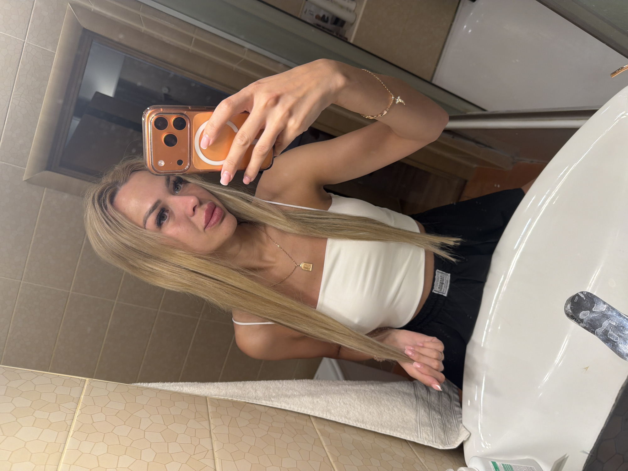A woman with blonde hair takes a selfie in a bathroom with beige tiled walls. She is wearing a white crop top and black shorts. She is holding an orange-cased smartphone in her right hand and looking into a mirror. A sink is visible in the foreground. The scene is set in Kyiv, Ukraine.