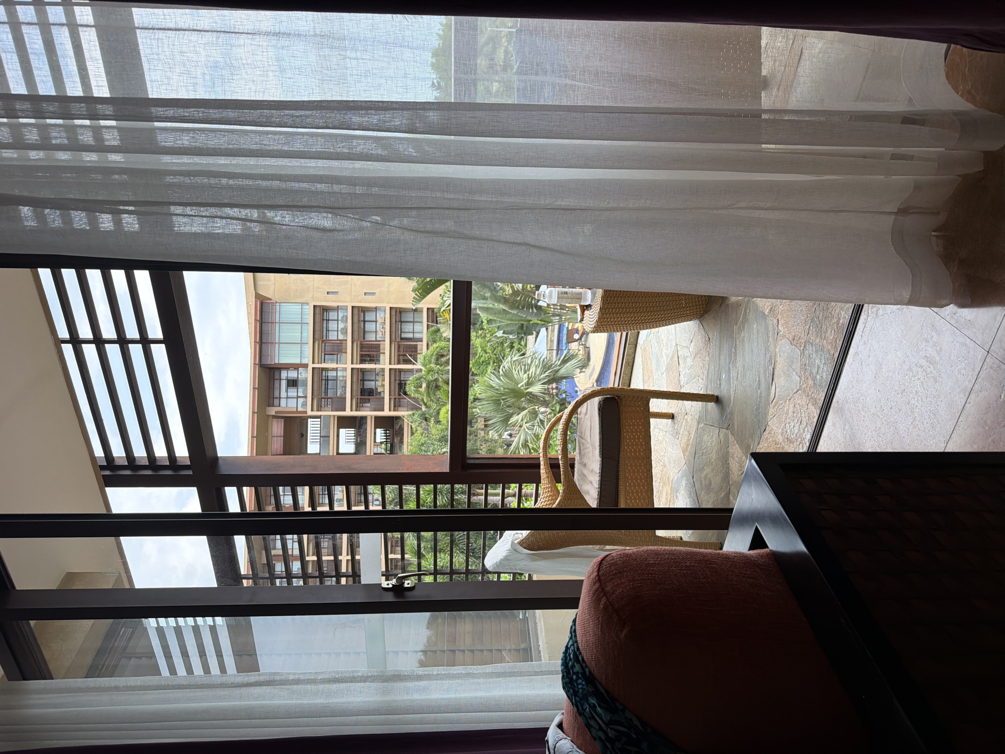 A hotel room view looking out onto a balcony and beyond to a resort-style courtyard with a pool. The room has a large glass sliding door, partially obscured by sheer white curtains. A dark wood frame separates the interior of the room from the balcony. In the foreground, a portion of a reddish-orange armchair is visible. On the balcony, a wicker chair with a cushioned seat and back is positioned. A small, round wicker table is next to the chair, holding a clear plastic bottle of water. Beyond the balcony, a multi-story building with balconies and dark-framed windows is visible, surrounded by lush green palm trees and other tropical foliage. A swimming pool with lounge chairs can be seen in the courtyard. The flooring on the balcony appears to be made of large, irregularly shaped stones. The sky is overcast.