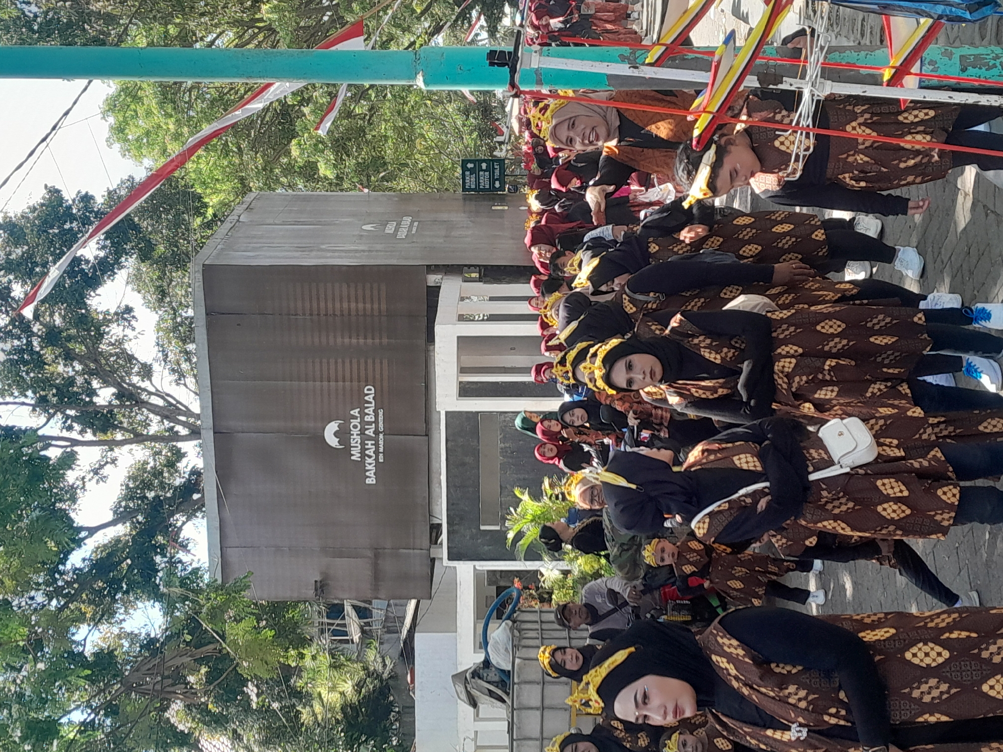 A group of people, mostly women and children, are gathered outdoors, possibly for a festival or parade. Many are wearing traditional Indonesian attire with brown and yellow batik patterns and golden headbands. In the background, a large, dark grey building displays the text "MUSHOLA BAKKAH AL BALAD" with an address below that reads "BITH MARON, GENTENG". The scene suggests a public gathering in the city of Genteng, Indonesia, with a festive or cultural event taking place. Red and white Indonesian flags are visible, strung across wires above the crowd.