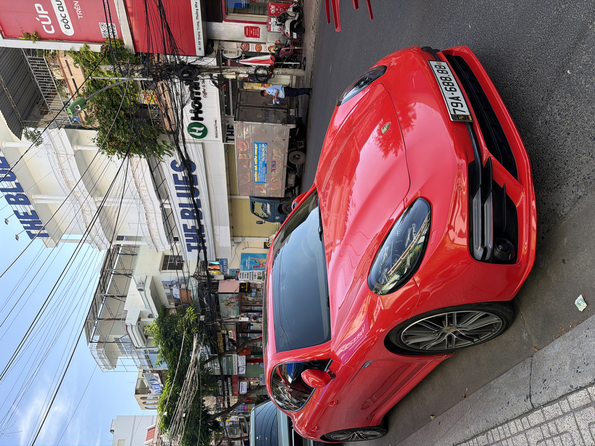 The image shows a bright red Porsche Panamera parked on a city street in Nha Trang, Vietnam, with its front end angled towards the camera. The car is parked adjacent to a sidewalk with a curb. In the background, there are multi-story buildings with various signage, including "THE BLUES" and a green "H Home Coffee & Tea" logo. A delivery truck is parked behind the Porsche, and a person in a light blue shirt and dark pants is standing next to it. There are numerous electrical wires suspended overhead, typical of Vietnamese city streets. A scooter is parked on the sidewalk near a red and white umbrella. The sky is partly cloudy. The license plate of the Porsche is visible and reads "79A-688.88".