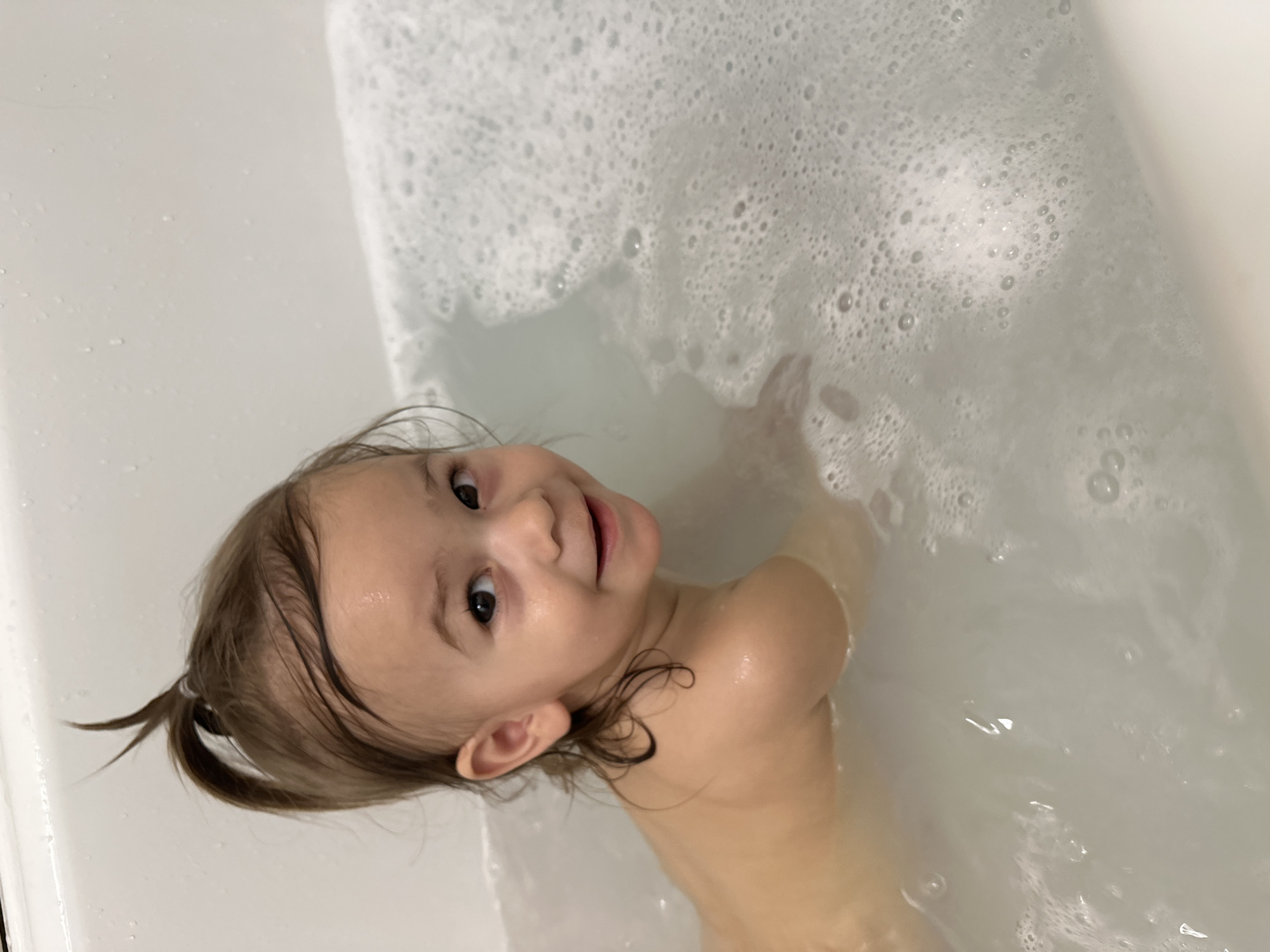 A young child is partially submerged in a white, bubbly bathtub, with their head and upper body visible. The child has light brown hair pulled back in a small ponytail, and is looking towards the camera with a slight smile. The bathtub is filled with water and soap bubbles, and the child's hand is visible near the bubbles. The scene is indoors, with the white surface of the bathtub dominating the background. The location cannot be determined as Kharnavy, Russia.