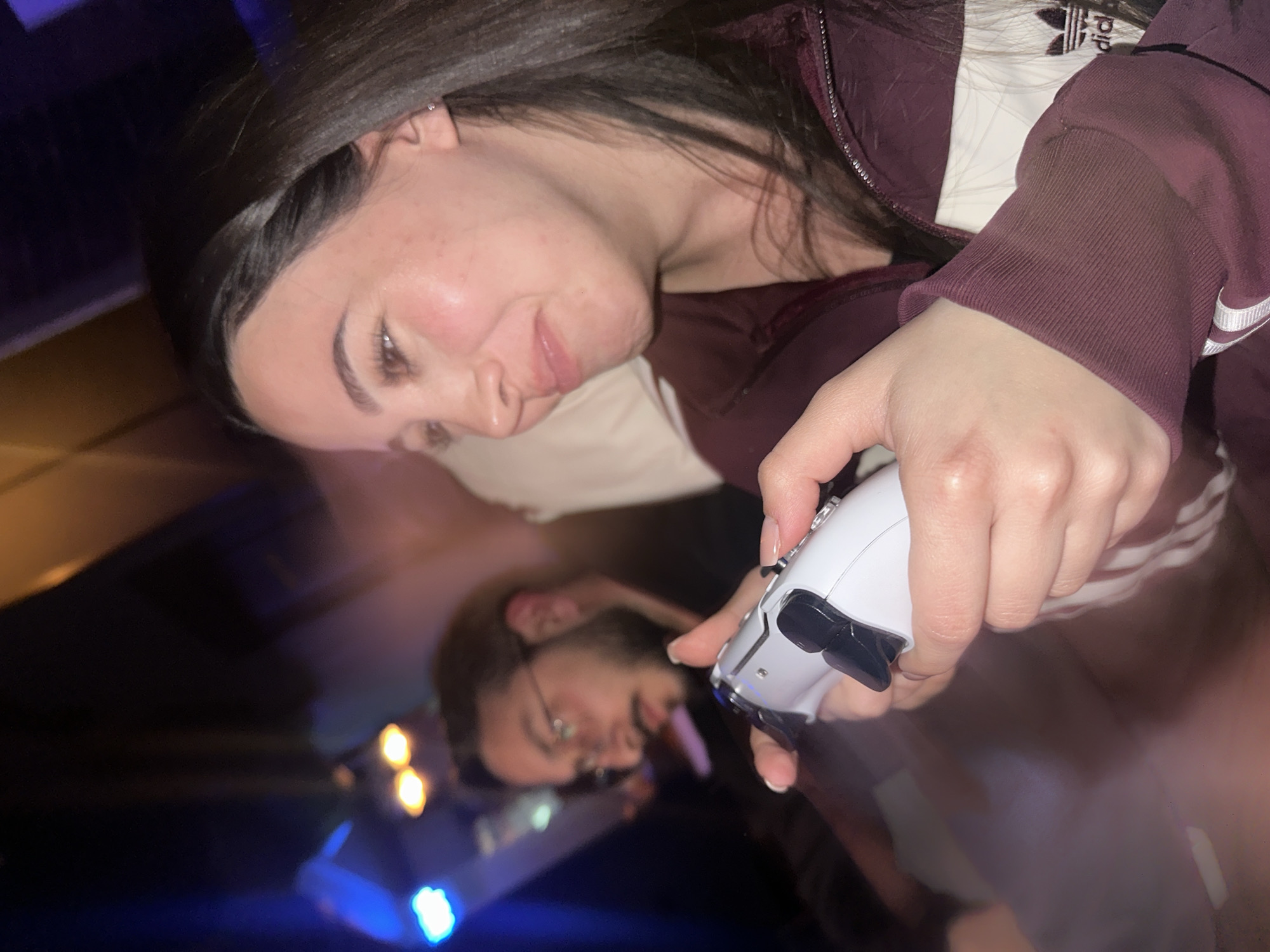 A blurry photograph shows a woman holding a white PlayStation 5 DualSense controller. She is wearing a maroon Adidas jacket with white stripes. In the background, partially visible, is another person with glasses. The lighting suggests an indoor setting, potentially a home or entertainment venue. The image is blurry with some light flares. There is no indication of the location in Yerevan, Armenia.