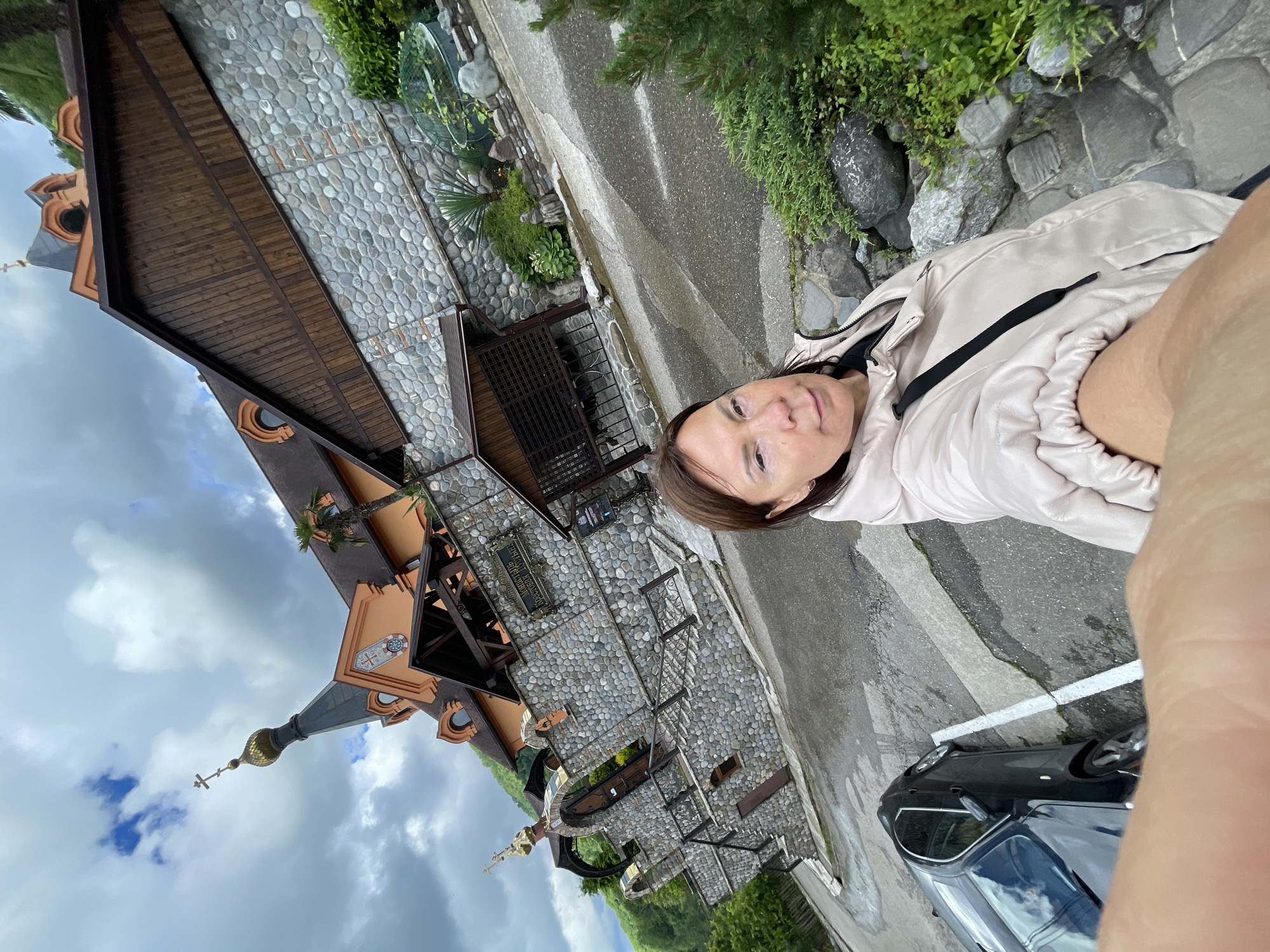 The image is a selfie taken by a woman wearing a light pink jacket and facing the camera. She is standing on a street, with a black car partially visible in the bottom right corner. Behind her is a building with a unique architectural style, featuring a stone facade, wooden elements, and a church-like spire with a golden dome and a cross. The sky is cloudy. The building appears to be located in Solokh-Aul, Russia.