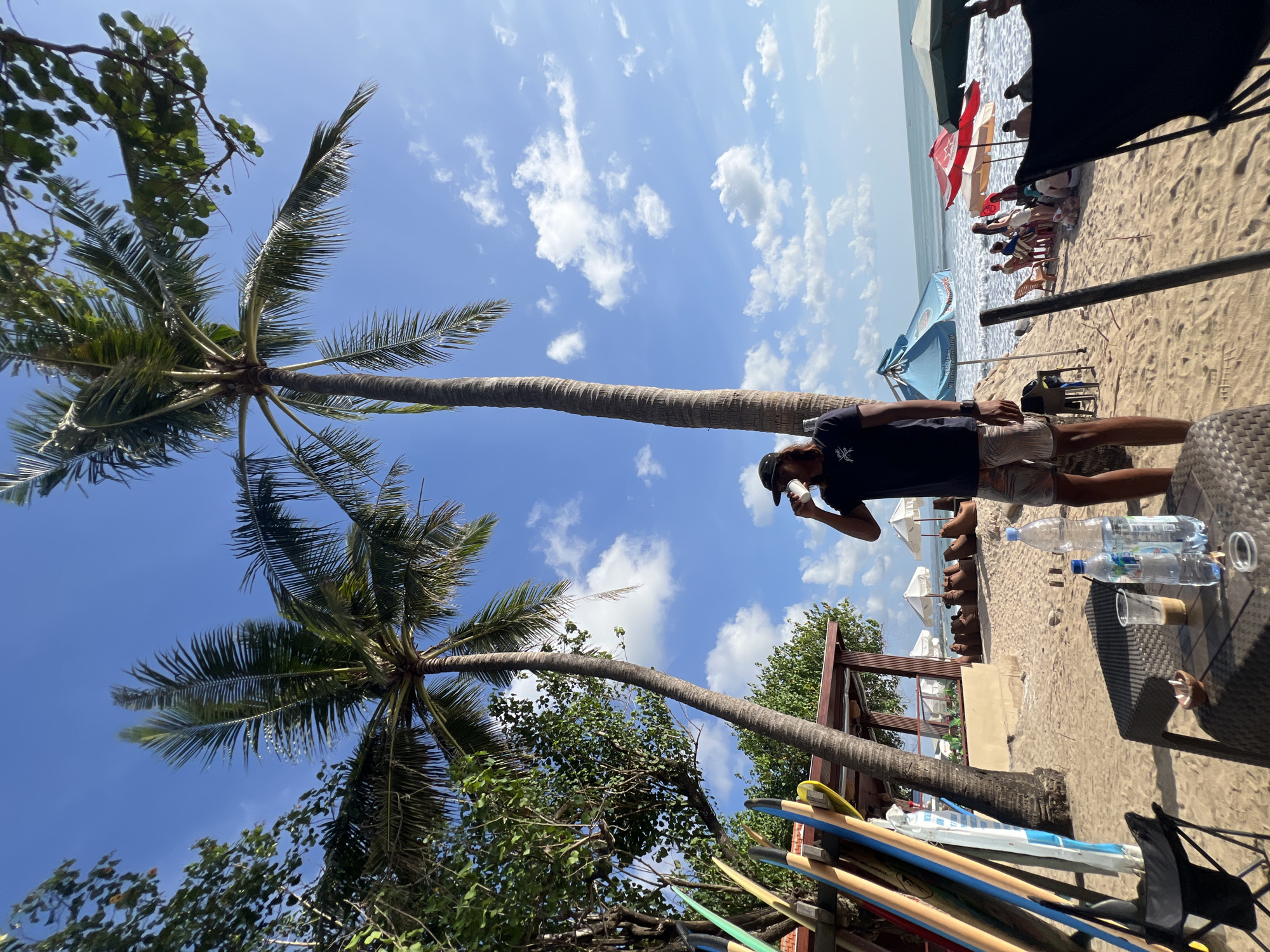 In Kuta, Indonesia, on a sunny day, a man wearing a dark t-shirt, patterned shorts, and a baseball cap stands on a sandy beach, holding a cup to his lips. The man is positioned between two tall palm trees, with their fronds reaching towards a bright blue sky dotted with white clouds. To the left of the man, a rack of colorful surfboards leans against a low structure. In the foreground, a woven table holds two bottles of water, a clear plastic cup with a drink, and an ashtray. Beyond the man, the beach stretches towards the ocean, where gentle waves are breaking. Several beach umbrellas and chairs are scattered across the sand, with people relaxing on them. The overall scene depicts a casual beachside setting in Kuta.