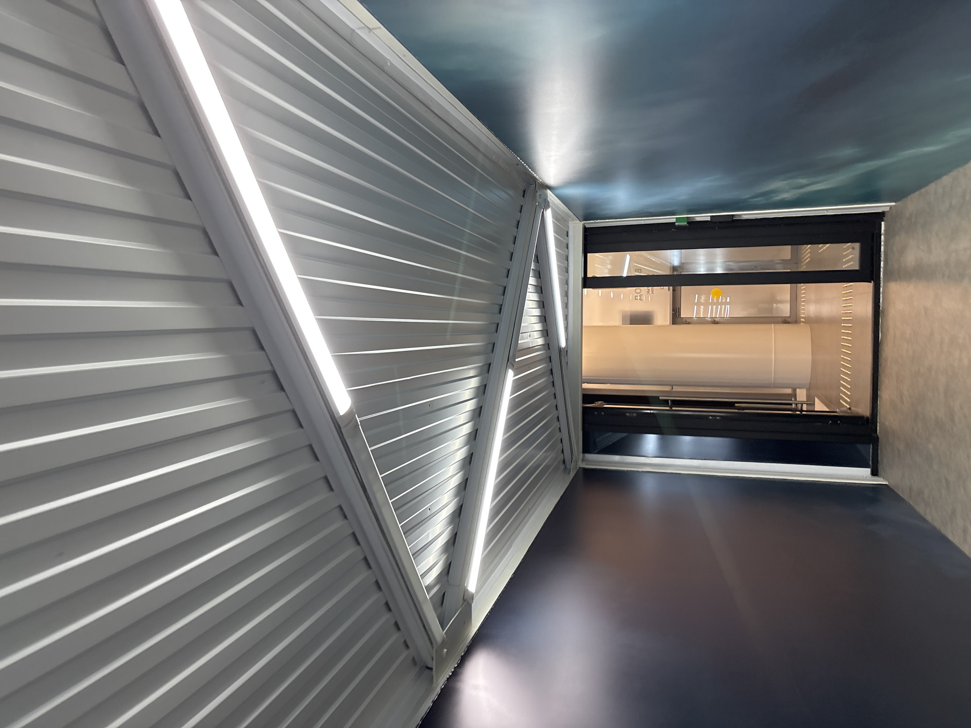 The image depicts the interior of a modern passageway. The left wall is composed of corrugated metal sheets with two diagonal white supports and integrated LED light strips. The ceiling is painted with a sky-like design. The floor is dark, reflective material. A window or opening is visible at the end of the passageway, revealing the interior of a building, showing what appears to be equipment and machinery. No people are visible in the scene. The scene appears to be located in the city of Новое Мартьяново, Russia.