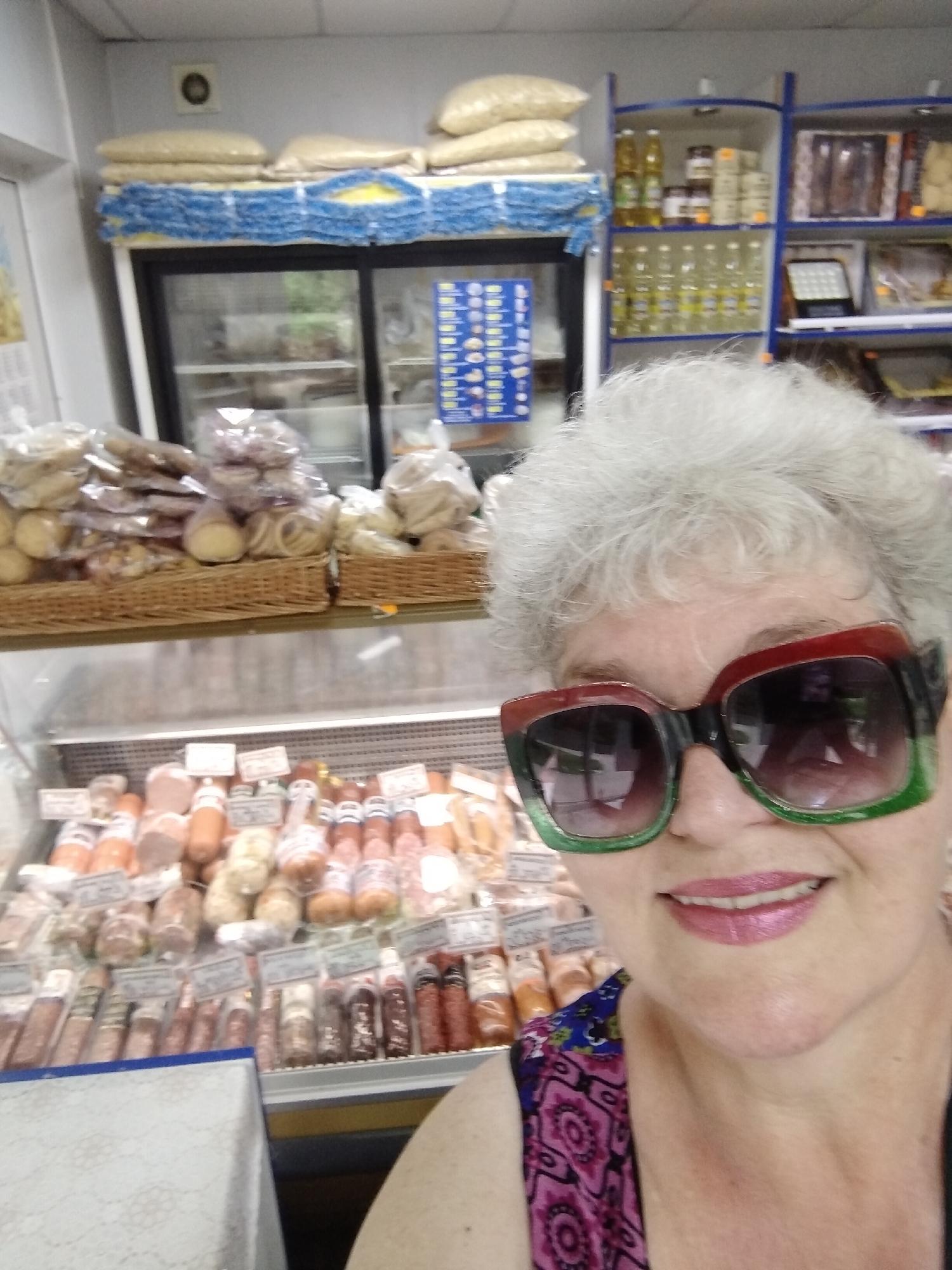 The image depicts a selfie taken by a woman inside a grocery store or market in the city of Horishni Plavni, Ukraine.
In the foreground, a woman with grey, curly hair is seen from the shoulders up. She is wearing oversized square-framed sunglasses with dark lenses, which are red on the top and green on the bottom. Her lips are painted with light pink lipstick, and she has a faint smile. She is wearing a sleeveless top featuring a blue and purple/pink geometric pattern.
Behind the woman, the interior of a retail food establishment is visible. A long, horizontal refrigerated display case occupies the lower mid-ground, filled with various types of sausages and cured meats, individually wrapped in plastic or netting and displaying small labels. To the left, a white counter or table with a textured surface is partially visible.
Above the refrigerated meat display, two wicker baskets hold numerous items packaged in clear plastic bags, appearing to be baked goods or snacks. Behind these baskets and to the left, a large, double-door refrigerator with glass doors is present, containing indistinct items. On top of this refrigerator, several large sacks of light-colored contents, possibly grains or nuts, are stacked. Below these sacks, a blue and yellow patterned fabric or material is layered.
To the right of the scene, a set of white and blue open shelving units are stocked with various products. The upper shelves display bottles of cooking oil with yellow liquid, jars, and packaged boxes. The lower shelves also contain multiple bottles of cooking oil. Further right, additional shelves with packaged goods are visible, and one shelf displays a dark, square object with multiple small, bright lights, possibly a compact light fixture or a specialized display. The walls of the establishment are white, and a dark circular object, possibly a vent or speaker, is mounted high on the wall in the upper left background.