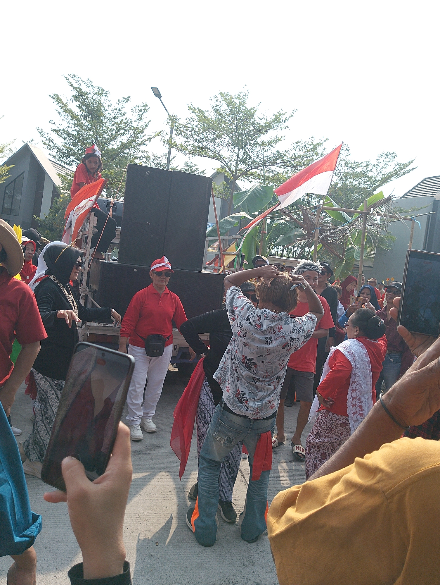 In the city of Karawang, Indonesia, a group of people are gathered outdoors, possibly for a cultural event or celebration. The scene is dominated by a truck equipped with a large speaker system, suggesting a performance or rally is underway. Several individuals are dressed in red and white, the colors of the Indonesian flag, which is prominently displayed, waving in the breeze. Some people are dancing, while others are observing. A young girl in a red top and a festive hat holds a small Indonesian flag. A person in the foreground is taking a photo with their smartphone. The background shows modern buildings and lush green trees, indicating a contemporary urban setting. The overall atmosphere appears festive and lively.
