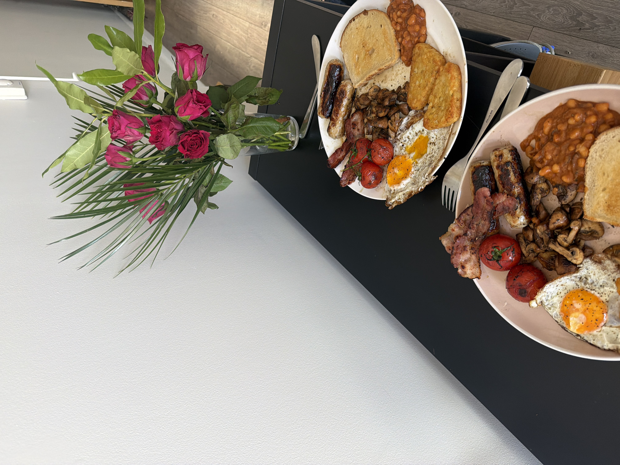 The image shows two plates of full English breakfast next to a vase of roses on a table. The plates contain fried eggs, sausages, bacon, toast, baked beans, grilled tomatoes, mushrooms, and hash browns. Forks are placed beside the plates. The table is partially white and partially black. The image does not provide information about the location in the city of London or any people.