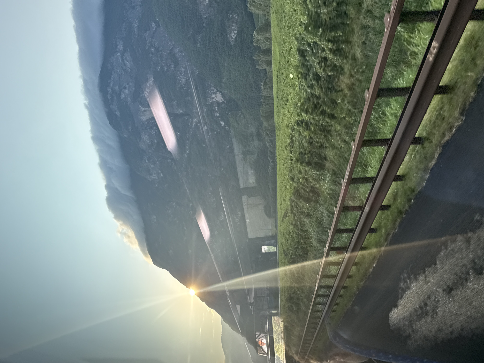 The image captures a mountainous landscape viewed from a moving vehicle, likely a train, approaching Mezzocorona, Italy. The sun is setting or rising behind a large, steep mountain, casting a golden glow and lens flare across the scene. A layer of wispy clouds is draped across the upper part of the mountain. In the foreground, a green field stretches out, bordered by a rusty metal guardrail and a dark asphalt road. The vehicle's interior is partially visible on the left, with a textured surface and what appears to be a seat. A billboard featuring a person is also visible on the left, attached to an overpass structure. The overall impression is one of a scenic journey through the Italian countryside.
