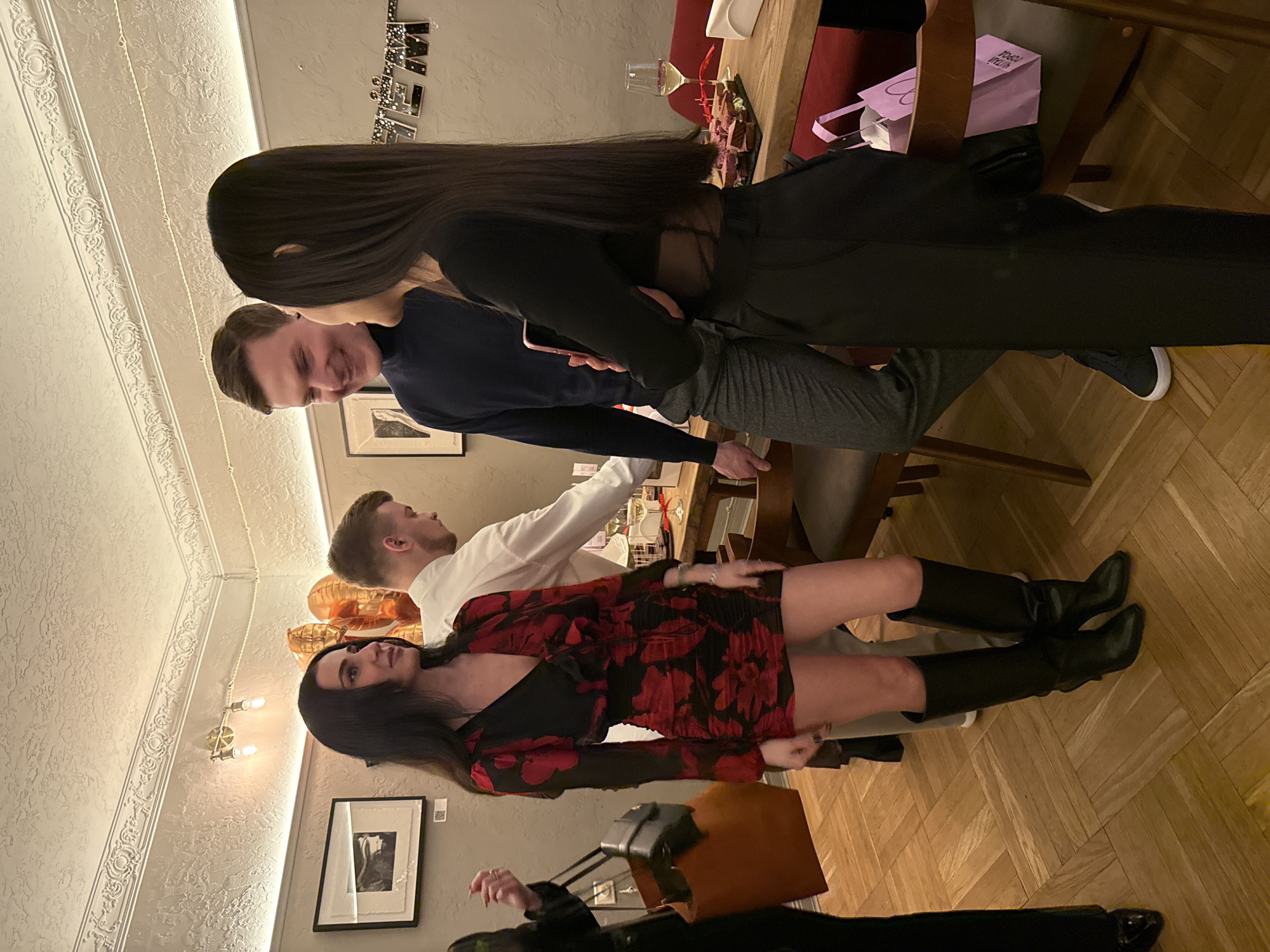 The scene depicts a group of people inside a room that could be located in a restaurant or a private event space in Moscow, Russia. The room has patterned walls and a herringbone wood floor. Four individuals are visible: a woman in a black and red floral dress and black boots, a man in a white shirt, a man in a navy blue shirt, and a woman with long black hair wearing a black top and pants. The people are standing near a table. There is food and wine on the table, along with a pink shopping bag. Another woman with a large brown purse is walking into the frame on the left side of the photo.