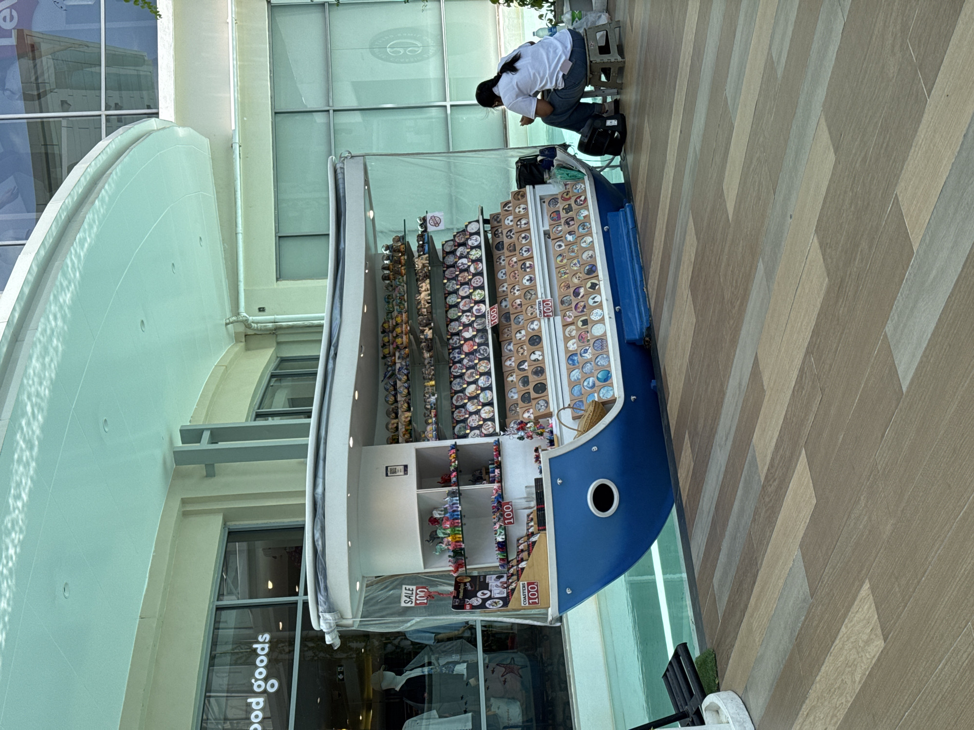 In Patong, Thailand, a woman sits on a small stool outside a souvenir boat-shaped stall. The stall displays numerous small figurines and round coasters with various designs. A "SALE 100" sign is visible. The stall is positioned next to a shallow pool of turquoise water, reflecting the overhead canopy. The ground is paved with wooden planks. In the background, the facade of a building features large windows and signage, including "good goods". The woman, wearing a white shirt and blue jeans, is looking down at her phone.
