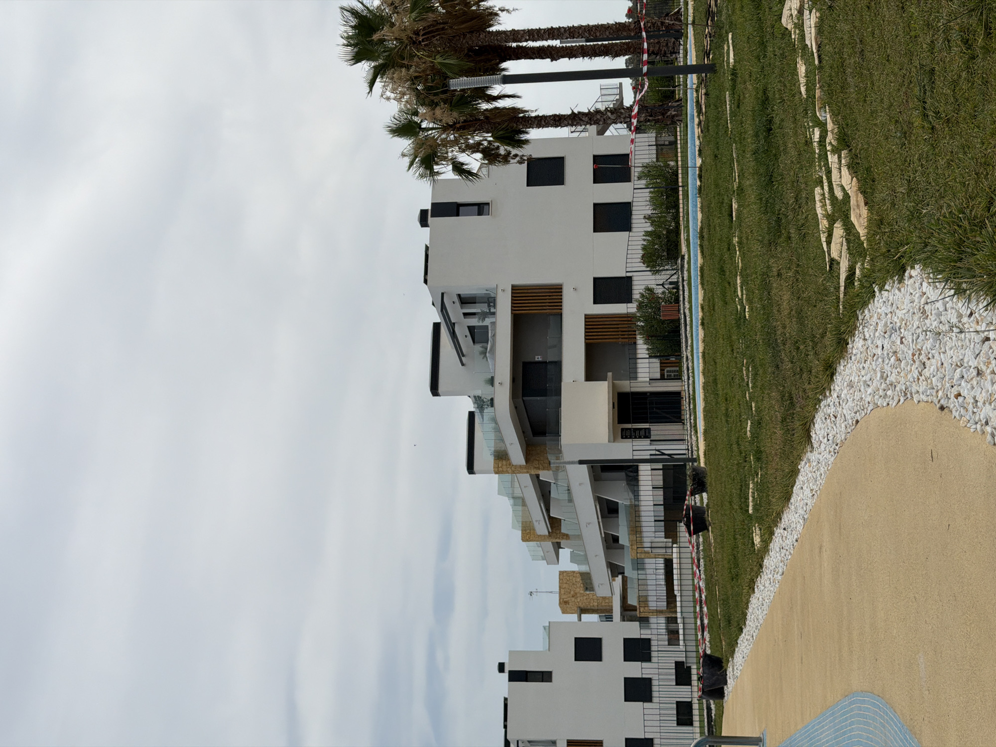 An exterior medium shot captures a modern residential building complex under an overcast sky in Finestrat, Spain. The white, multi-story buildings feature clean lines, dark-framed windows, and balconies with glass railings and wooden accents. Palm trees stand to the right of the frame. A pathway made of light-colored aggregate borders a grassy area, which is interspersed with flat stones. A swimming pool with blue mosaic tiles is visible in the background, behind a black metal fence. Red and white caution tape is strung between posts and objects on the grass, suggesting ongoing construction or maintenance. Two dark, cylindrical containers are placed near the fence. The scene is devoid of human activity.