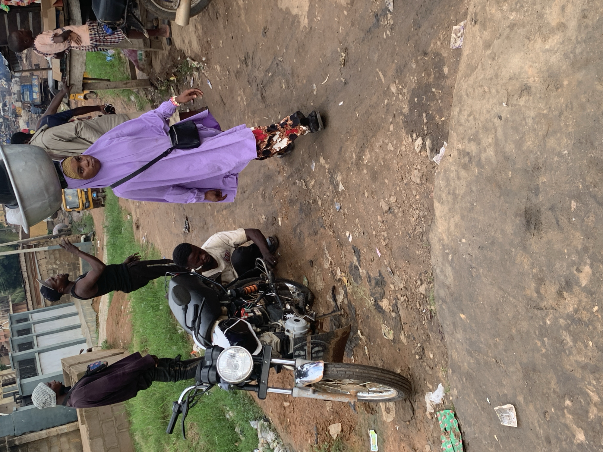 In Shagamu, Nigeria, a woman in a purple hijab and patterned pants walks with a large metal bowl balanced on her head, a black purse slung across her body. To her left, a man in a dark shirt and beanie stands beside a motorcycle. To the right of the woman, a child in a pink shirt and checkered shorts stands near a market stall. In the foreground, another man kneels beside the motorcycle, appearing to be working on it. The scene unfolds on a dusty, unpaved road, indicative of a bustling urban environment.