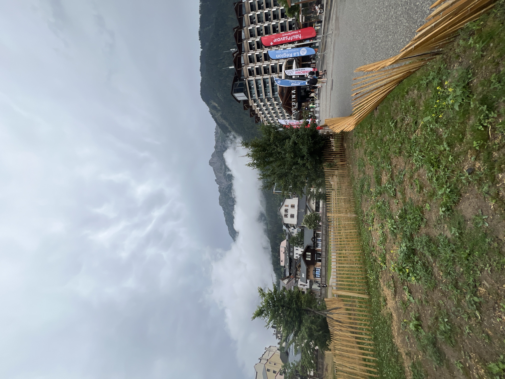 This is an outdoor, eye-level, wide-angle shot of a street scene in Chamonix-Mont-Blanc, France, on a cloudy day. In the foreground, a wooden fence encloses a patch of dry grass and sparse vegetation. Beyond the fence, a paved road runs diagonally to the right. Several people are walking on the road, which leads to a large, multi-story hotel with a sign that reads "ALPINA." In front of the hotel, numerous tall, colorful flags are planted. To the left of the hotel, a cluster of buildings, typical of mountain towns, are visible, with a mountain range looming in the background, partially obscured by clouds.