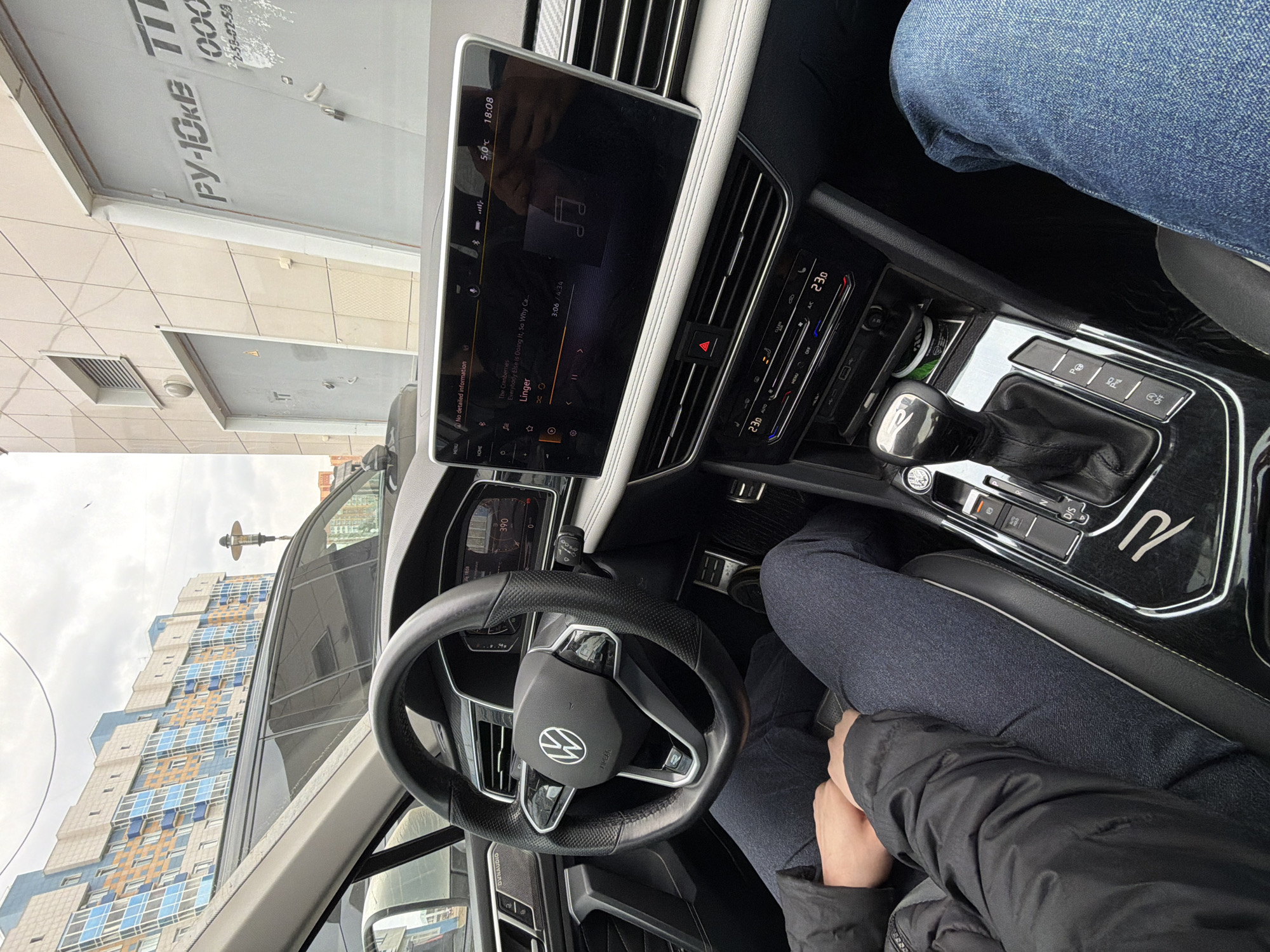 Here is a description of the image:
The image shows the interior of a car, focusing on the dashboard and the view through the windshield. The car appears to be parked near a building with a sign in Russian (“РУ-OKE T 1000”). The infotainment system displays a song titled "Linger" by The Cranberries. The car's climate control is set to 23 degrees Celsius. The image captures a person’s legs and hands inside the vehicle. The angle is tilted, showing a high-rise building through the windshield. Based on the building's architecture visible through the windshield, and the style of the Russian signage, the location is likely within the city of Krasnoyarsk, Russia.