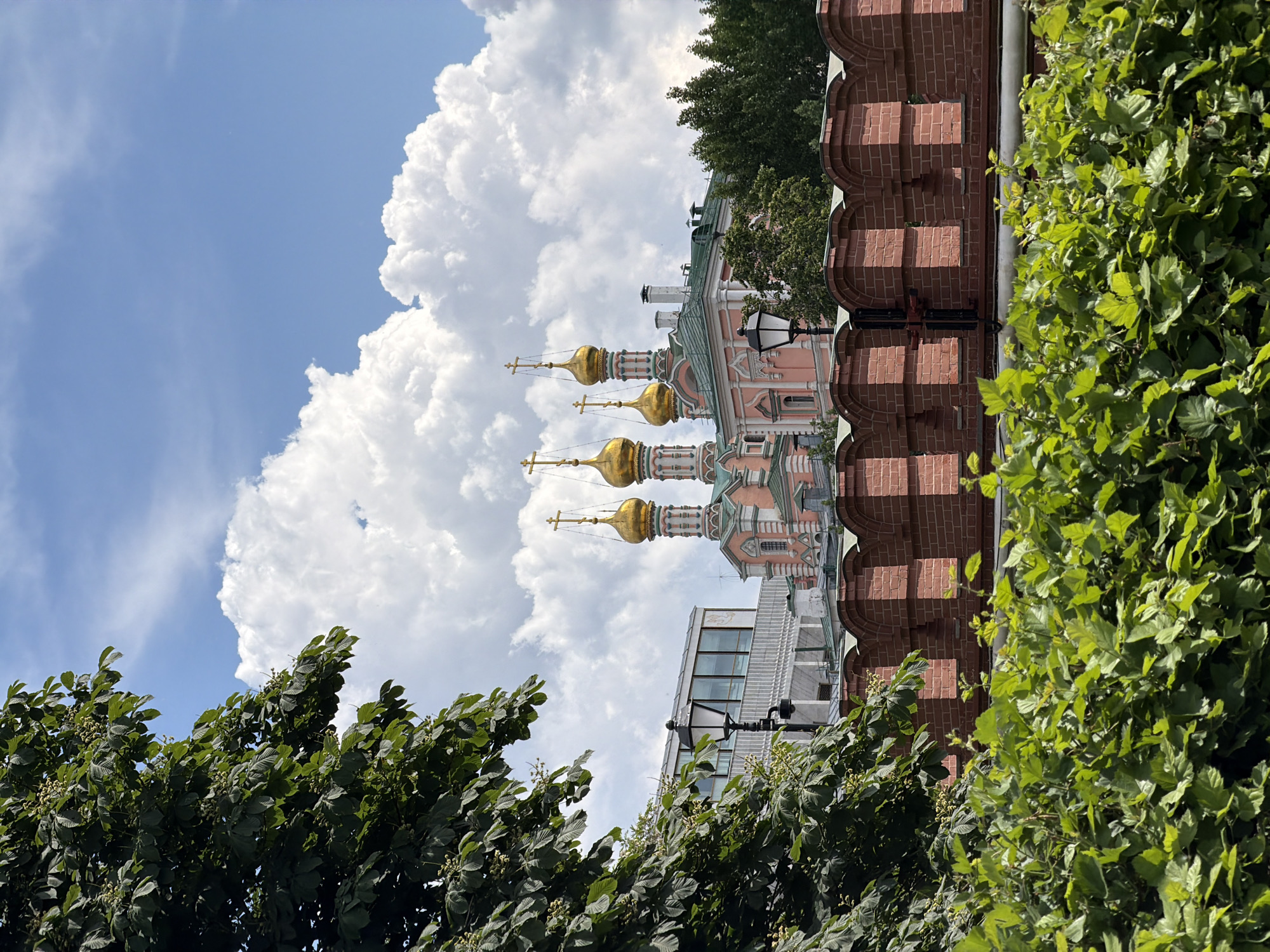 An outdoor, brightly lit image captures a picturesque scene centered around an ornate Eastern Orthodox church. The church building, rendered in a distinctive light pink or salmon color, features multiple gleaming golden onion domes, each topped with a cross. Intricate architectural details are visible on its facade and domes.
To the right of the church, a sturdy red brick wall with a unique, decorative scalloped top edge frames a portion of the scene. Lush, vibrant green foliage from trees and bushes densely occupies the foreground, extending across the bottom and partially up the left side, slightly obscuring the view of the structures. A traditional black street lamp stands near the church and brick wall, adding to the urban aesthetic. In the background, partially visible behind the church, is a modern building characterized by its sleek design and glass windows, creating a contrast between historical and contemporary architecture.
The sky above is a clear, bright blue, dotted with large, fluffy white cumulus clouds, indicating fair weather and sunshine. The scene is devoid of people and any discernible activity, presenting a static, architectural landscape. No visible text is present. The time of day is clearly daytime, illuminated by bright natural light. The golden onion domes and architectural style strongly suggest a location in Eastern Europe, likely Russia.