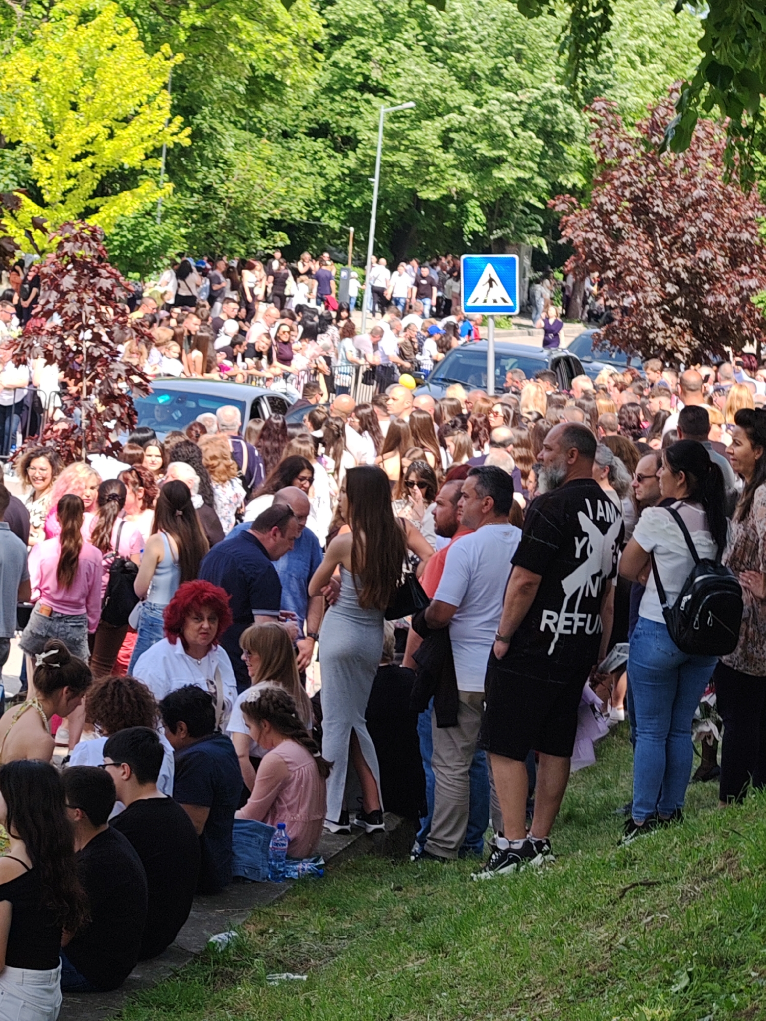 The image shows a large crowd of people gathered in a public area, possibly a park or square. The location is Shumen, Bulgaria as indicated by the license plate of the automobile.
**People:**
- A diverse group of people of various ages and ethnicities.
- Individuals are standing, sitting on the ground, or walking.
- Their attire varies, suggesting a casual setting.
**Objects:**
- Trees and greenery are visible in the background, creating a natural backdrop.
- A road sign with a pedestrian crossing symbol is prominently displayed.
- Cars are parked nearby.
- A water bottle is on the ground near seated individuals.
**Actions and Interactions:**
- People are mingling and interacting with each other.
- Some individuals appear to be observing an event or object of interest out of frame.
**Environment:**
- It appears to be a sunny day, and the lighting is bright.
- The setting is likely a public space in the city, possibly near a road.