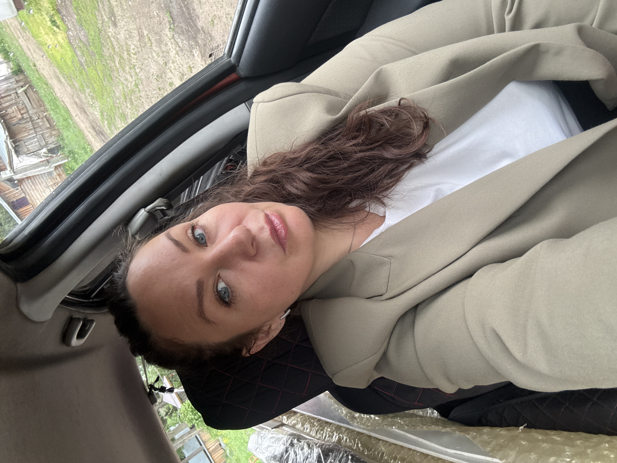 A woman with fair skin, blue eyes, and brown, curly hair is pictured from a high-angle perspective, likely taken from inside a vehicle. She wears a light-colored blazer over a white shirt. The vehicle's interior is visible, including the dashboard and window frame. Outside the window, a rural landscape with wooden structures and greenery can be seen. The location is Chaikovsky, Russia, based on contextual information. Other objects inside the vehicle include a black quilted seat cover and some plastic-wrapped items.