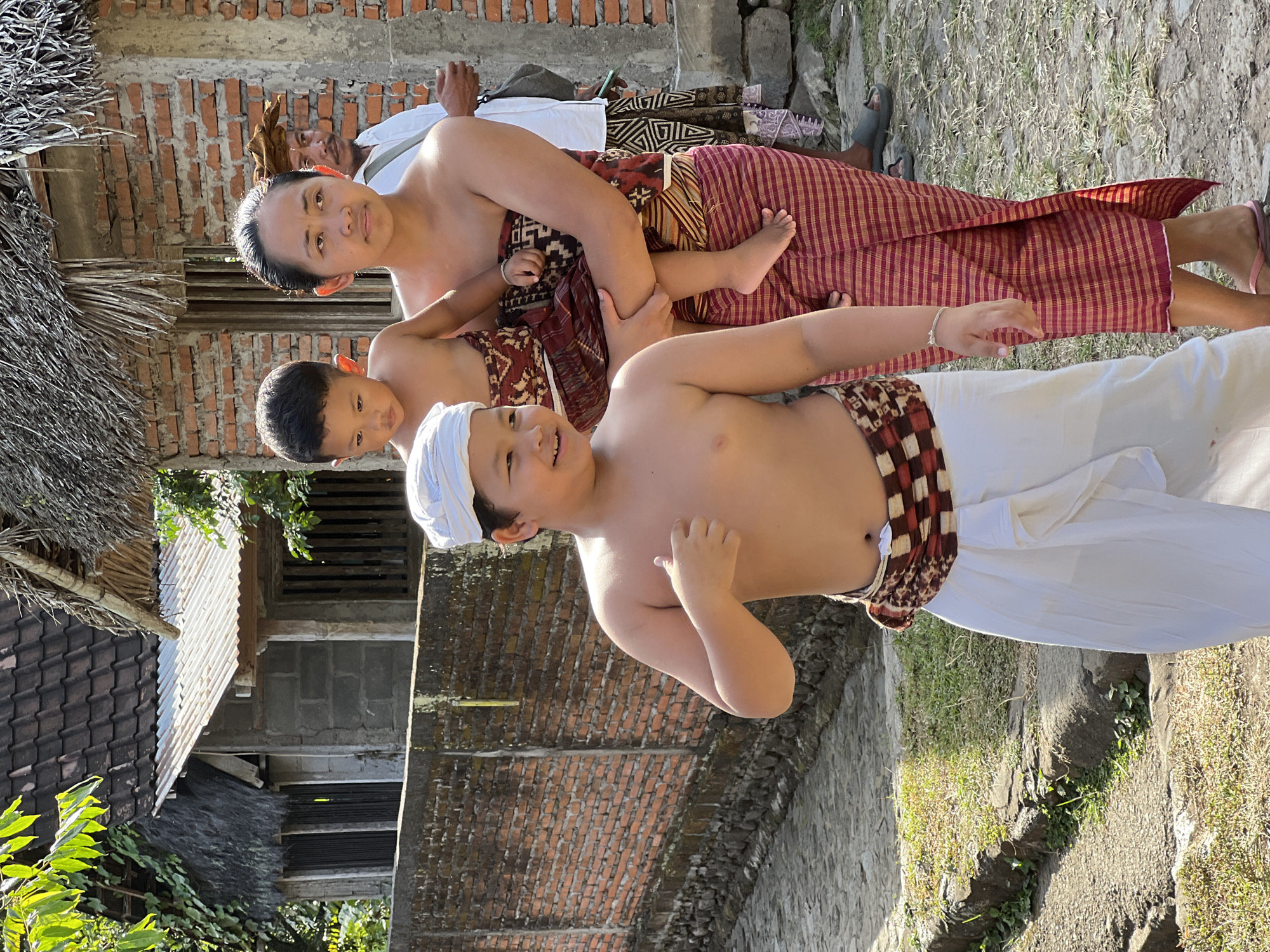 In Manggis, Indonesia, a young boy with fair skin, brown hair, and a white head covering stands in the foreground, giving a thumbs-up. He wears a white sarong and a patterned sash around his waist. To his right, a woman with dark hair carries a young child who is looking down. The woman wears a red and gold patterned sarong. Behind them, a man with dark hair wears a white shirt and a brown head covering. The scene takes place outdoors on a paved path, with a brick wall and buildings in the background. The sun is shining brightly, casting shadows.