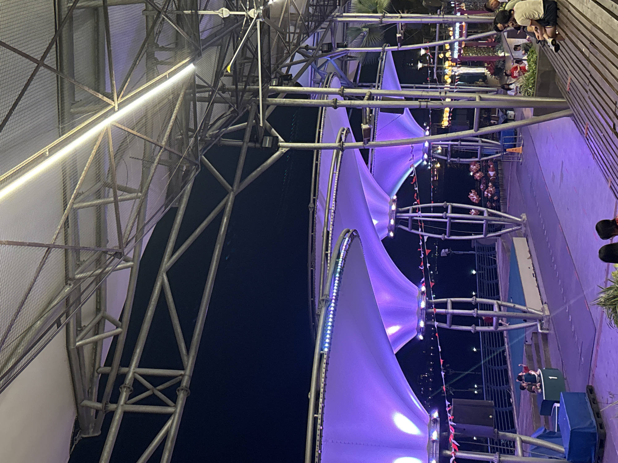 The image depicts an outdoor evening scene at the Ancol Beach City promenade in North Jakarta, Indonesia. The foreground features a wooden bench with several individuals seated, appearing to be engaged in conversation or observing the surroundings. Behind them, a paved area is illuminated by purple and white lights, highlighting a series of large, tent-like structures supported by metallic scaffolding. These structures are adorned with colorful LED lights along their edges.
Further back, a railing separates the promenade from a body of water, with lights visible on the opposite shore, suggesting a cityscape in the distance. Several people are scattered throughout the scene, some sitting on raised platforms and others walking. In the mid-ground, there's a stage-like area with a blue surface and steps. The overall atmosphere is festive, with the lighting creating a vibrant and modern ambiance.