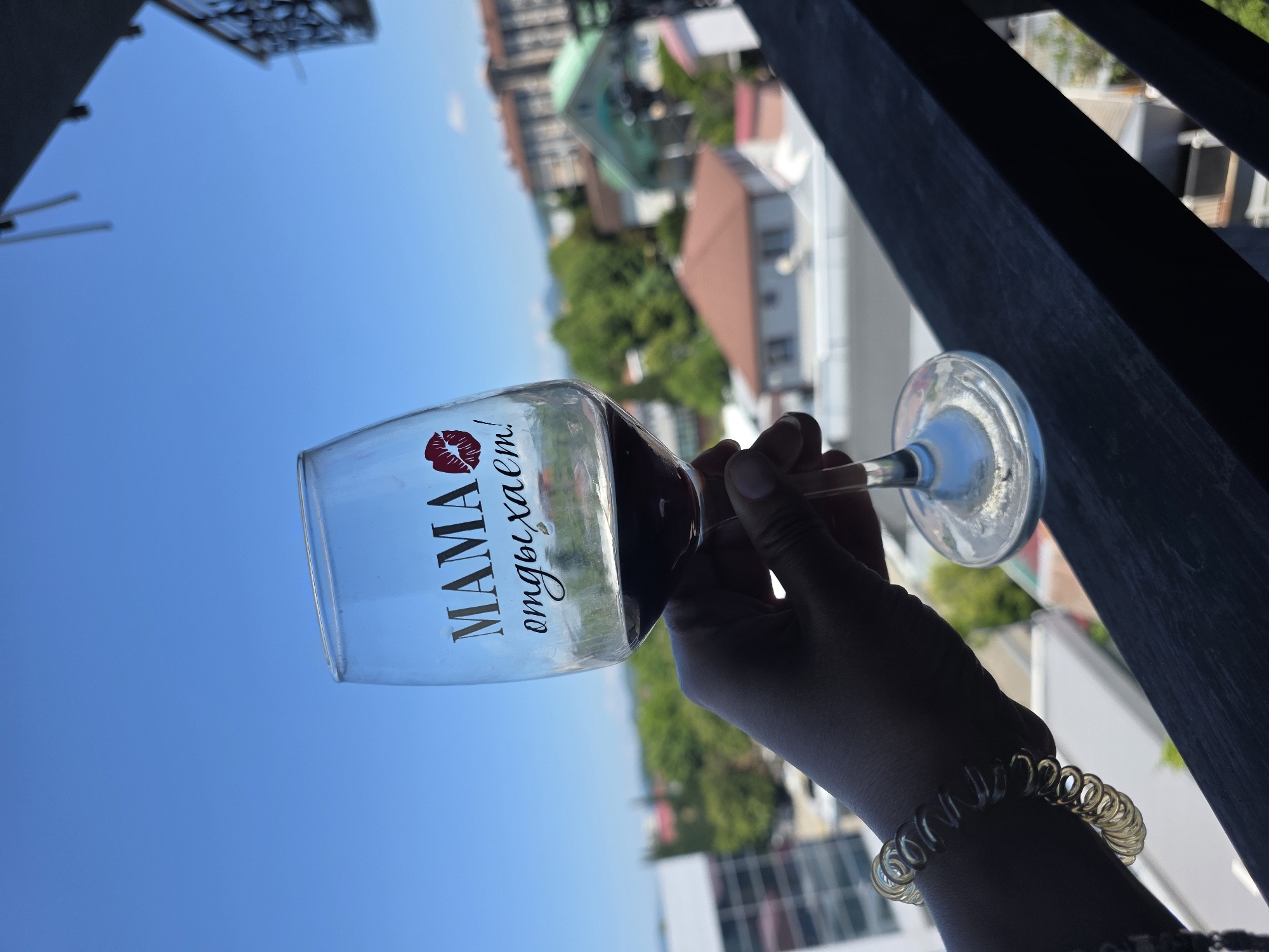 A hand holding a wine glass with red wine against a bright blue sky. The glass has the words "MAMA отдыхает!" and a red lipstick kiss mark printed on it. In the background, a blurry cityscape with buildings and trees is visible, suggesting an urban or suburban environment. The railing in the foreground indicates a balcony or terrace setting. The overall scene appears to be a moment of relaxation, possibly on vacation, in Sochi, Russia, given the architectural style and vegetation commonly found there.