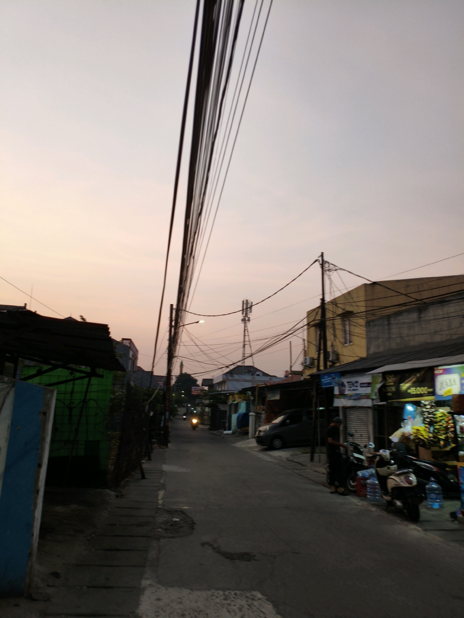 The image depicts a street scene at dusk in Bekasi, Indonesia. The sky is a pale orange and pink gradient, suggesting the sun has recently set. A dense network of overhead power lines crisscrosses the frame, supported by poles on either side of the street. On the left, a low-rise building with a dark, sloped roof is partially visible, its green wall weathered and aged. Further down the street on the right, a row of shops with awnings and illuminated signs line the road. A dark-colored car is parked in front of one of the shops. Two motorcycles are also present, one parked and another with a rider. A person, dressed in dark clothing, stands near a motorcycle, possibly interacting with someone or arranging goods. In the distance, a cell tower is visible against the sky. The road surface appears to be asphalt, with some patches of wear and tear. The overall impression is of a bustling, everyday street in a residential or commercial area of the city during the transition from day to night.
