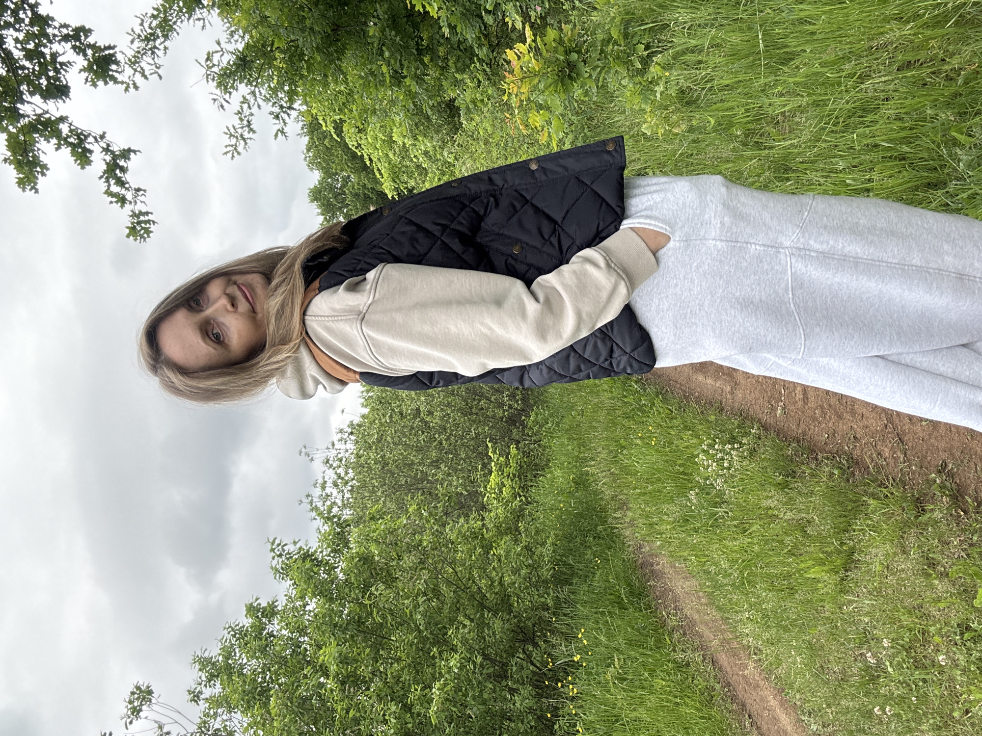 An adult female is depicted standing on a narrow dirt path in a natural outdoor setting, identified as Noviy, Russia. She is positioned slightly to the right of the frame, facing the camera with a subtle smile and her head tilted slightly to her left. Her long, light brown hair is parted on the side and falls over her shoulders. She is dressed in a light-colored hooded sweatshirt, potentially beige or light tan, layered with a dark, possibly black, quilted vest featuring brown shoulder patches and brass-toned snap buttons down the front. Light grey sweatpants cover her lower body, and her left hand is inserted into a pocket.
The environment consists of a verdant landscape. A well-trodden, unpaved path diagonally traverses the image from the lower left towards the upper middle, bordered on both sides by dense green grass and various broadleaf plants. Clusters of small yellow wildflowers are visible among the grass alongside the path. Beyond the immediate foreground, a dense growth of green bushes and trees forms a natural backdrop, extending into the mid-ground and background. The sky above is completely overcast with light grey clouds, indicating a cloudy or overcast weather condition without direct sunlight. No other individuals or man-made structures are discernible within the frame.