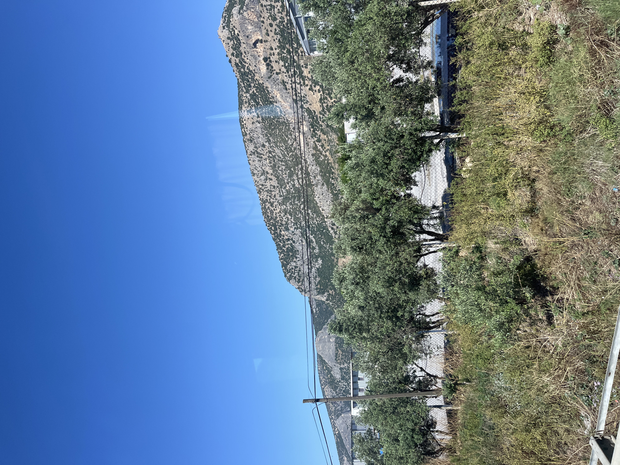 The image displays a bright, sunny landscape, likely captured in Torbalı, Turkey, and presented rotated 90 degrees counter-clockwise. On the left side, a vast and clear deep blue sky occupies a significant portion of the frame. A large, rugged mountain range, characterized by rocky slopes and sparse green and brown scrub vegetation, extends diagonally from the center towards the top right.
In the middle ground, a horizontal line of dense, dark green trees, possibly olive trees, runs across the frame, casting shadows. Behind these trees, a chain-link fence is visible, with hints of light-colored buildings or structures nestled at the mountain's base. The lower right portion of the image features a patch of mixed vegetation, including dry, yellowish grasses and greener, bushy plants. A utility pole, made of wood or concrete, with several power lines strung across it, cuts through the mid-ground, extending diagonally.
The scene is bathed in strong, direct sunlight, indicating midday or early afternoon conditions. The weather is clear and dry, typical of a Mediterranean climate. No people or visible text are present, contributing to a tranquil and natural atmosphere, despite the visible elements of human infrastructure like the fence, buildings, and power lines.