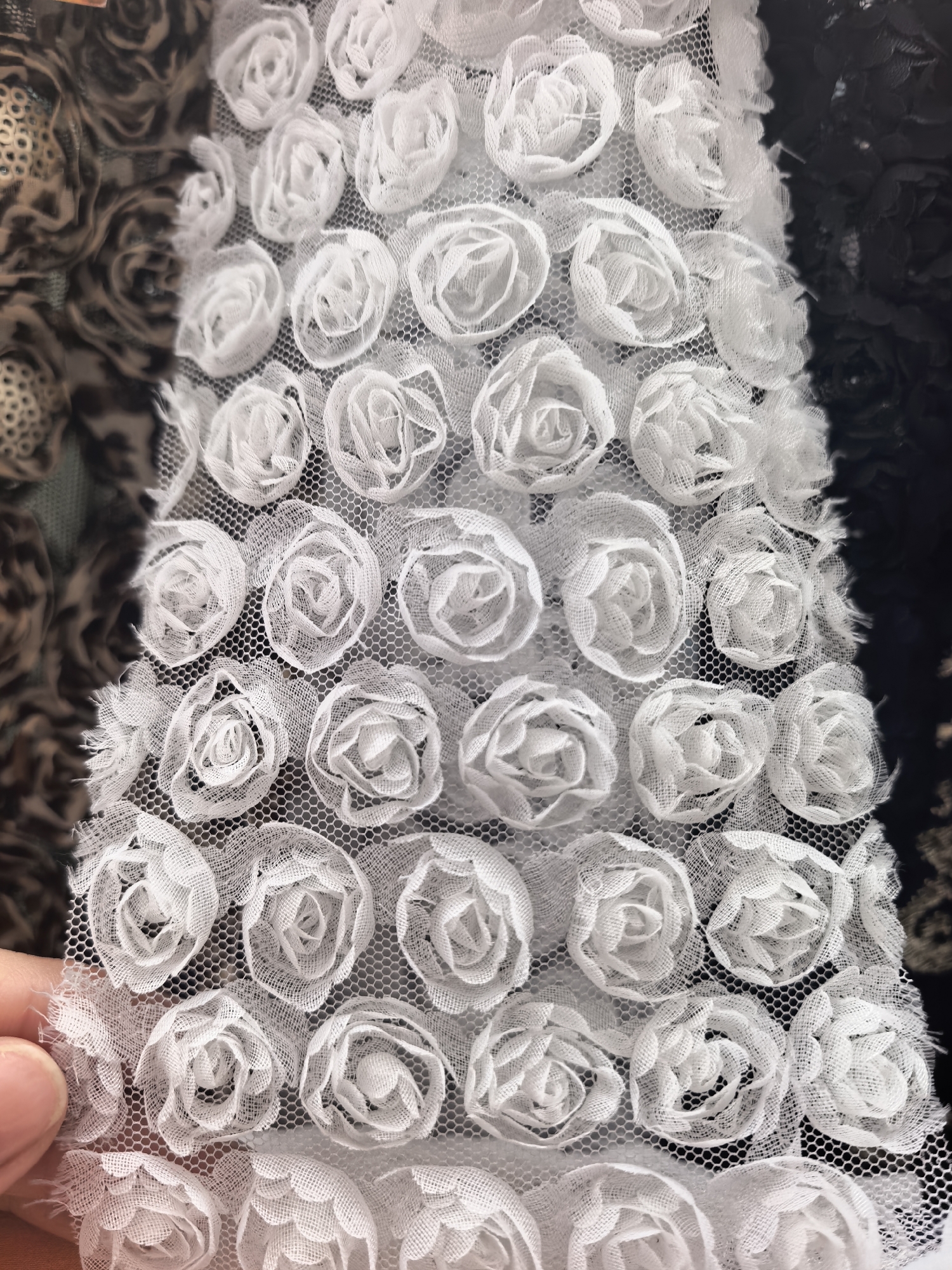The image shows a close-up of white fabric with small, rose-shaped embellishments arranged in a grid pattern on a net backing. A person's finger is visible holding the fabric at the bottom left corner. The background shows blurry fabric with a black and brown pattern. The location is unknown and cannot be determined from the image. Sterlitamak, Russia, is irrelevant to the content of the image.