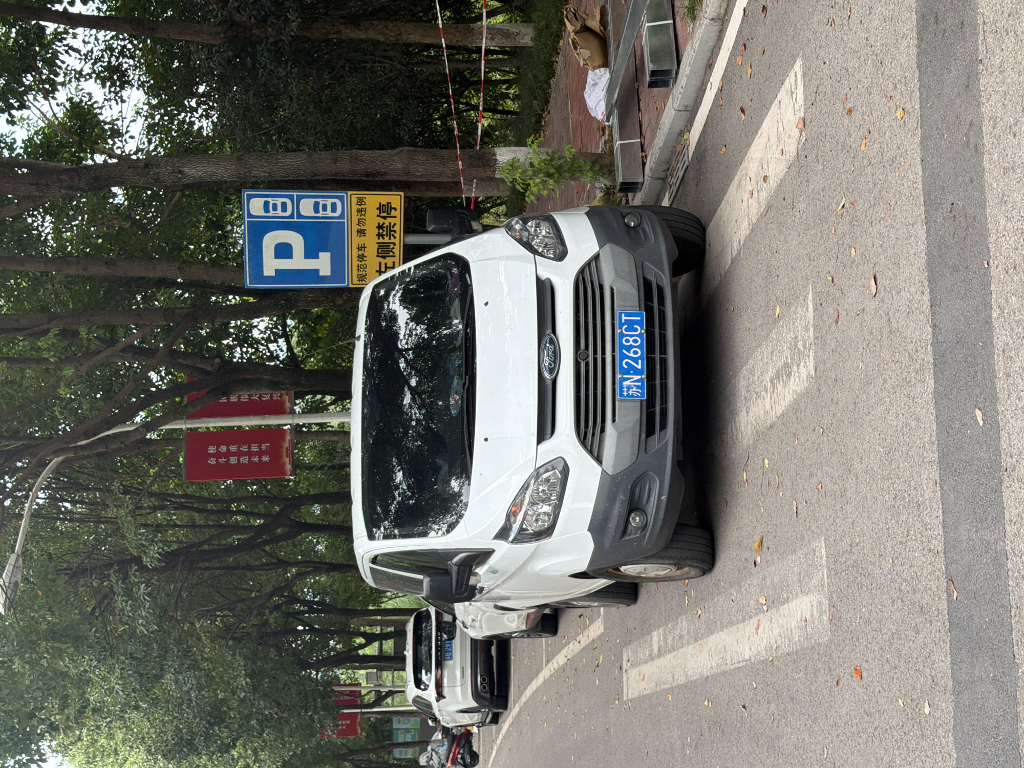 In Jiangjin, China, a white Ford Transit van is parked on a pedestrian crossing on a city street. The van's license plate reads "苏N·268CT". A white SUV is parked behind it. The street is lined with trees, and a blue and yellow parking sign is visible, indicating that parking is prohibited on the left. Red banners are hung on poles between the trees. Construction materials are stacked on the sidewalk next to the street.